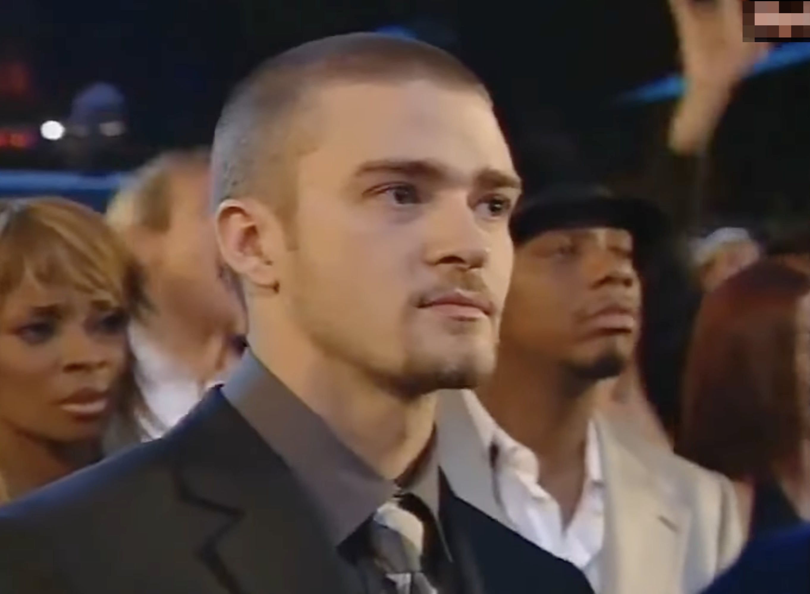 A person stands among others at an event, wearing a suit and tie, looking pensive
