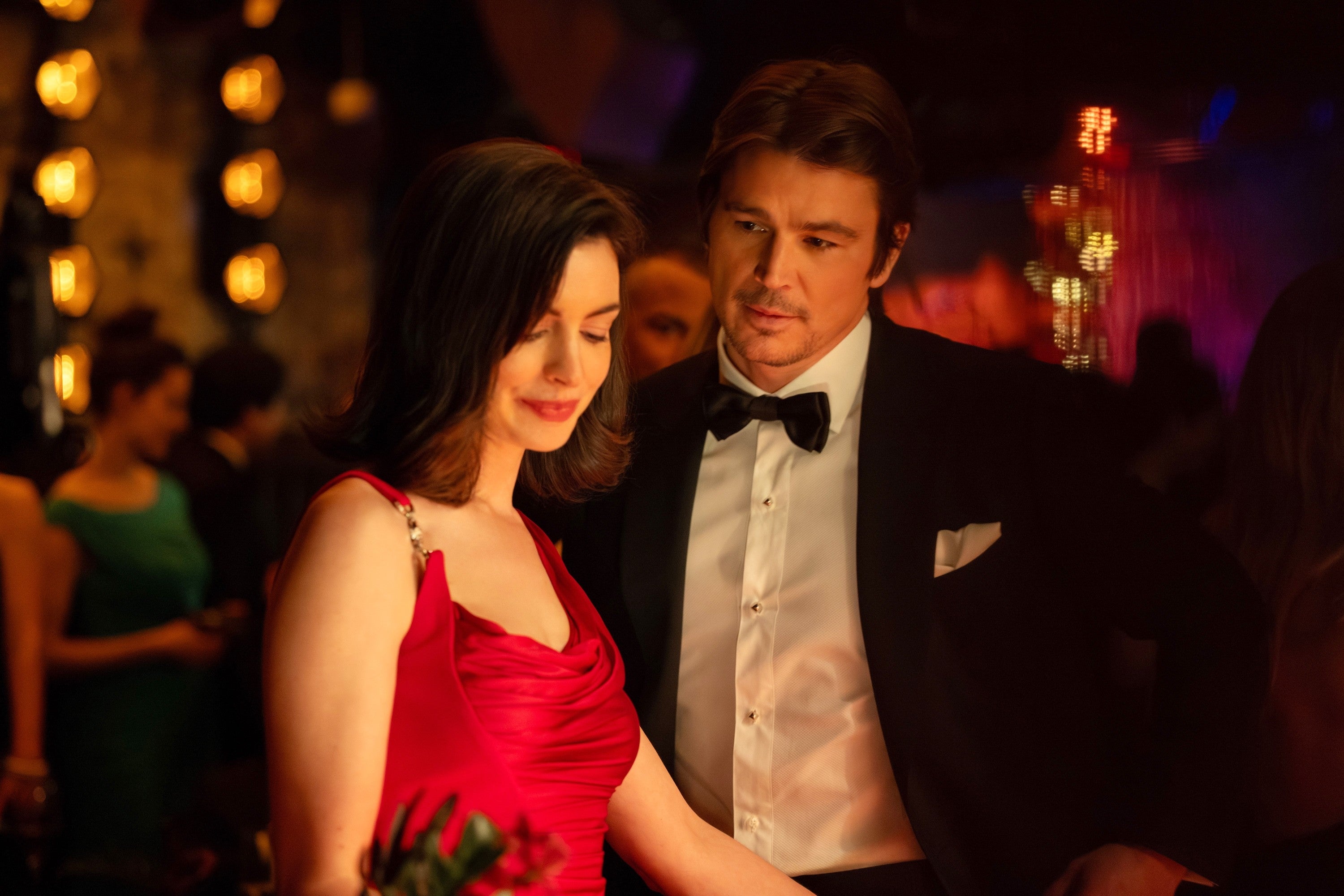 A couple in elegant attire shares a moment at a formal event; the woman wears a stylish red gown, and the man dons a classic tuxedo