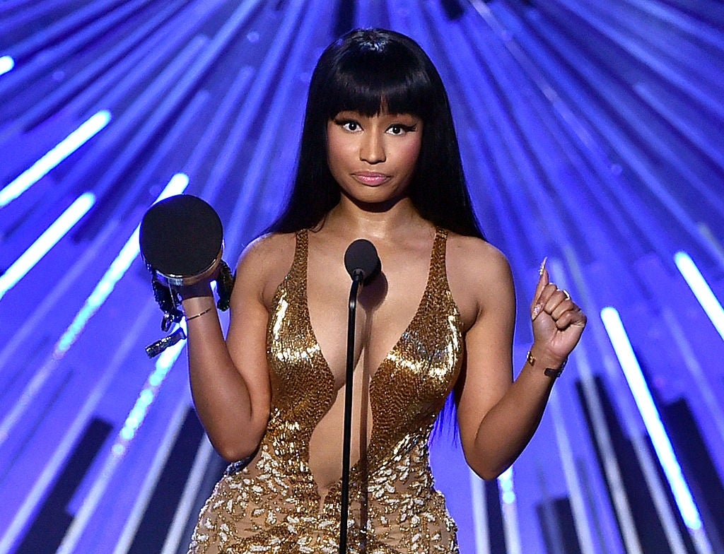 Celebrity in a shimmering, plunging gown holds an award on stage, standing before a fan-like backdrop, expressing a confident pose
