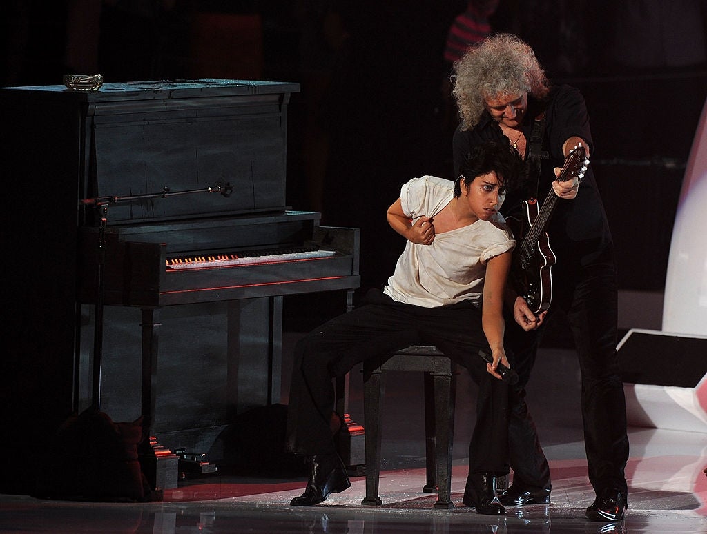 A performer in a white shirt and black pants sits at a piano, energetically engaging with a guitarist playing nearby on stage