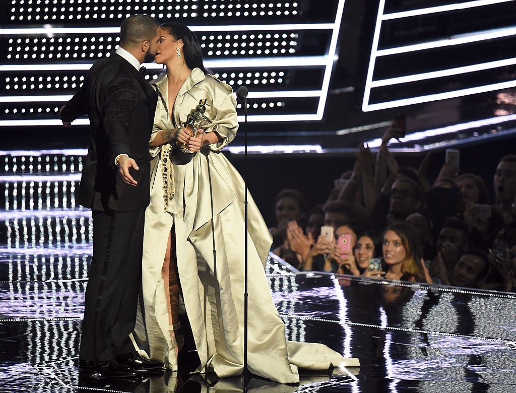 Two celebrities share a moment on stage at an award show. One wears a long, elegant coat and holds a trophy, while an audience watches