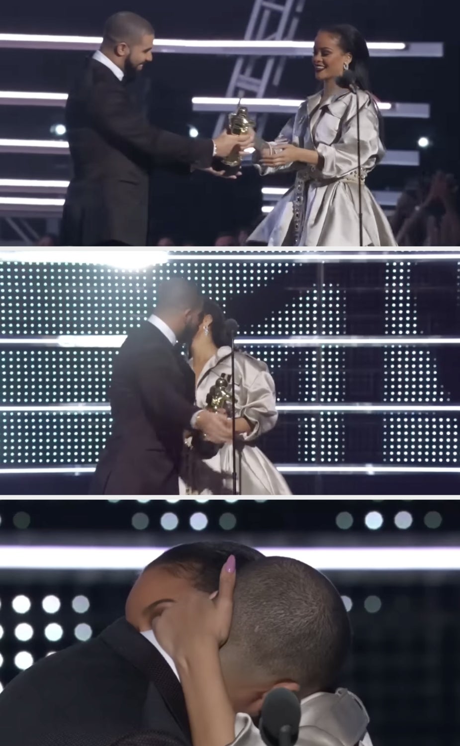 Two individuals on stage; one presents an award to the other, who is wearing an elegant gown. They share a warm embrace