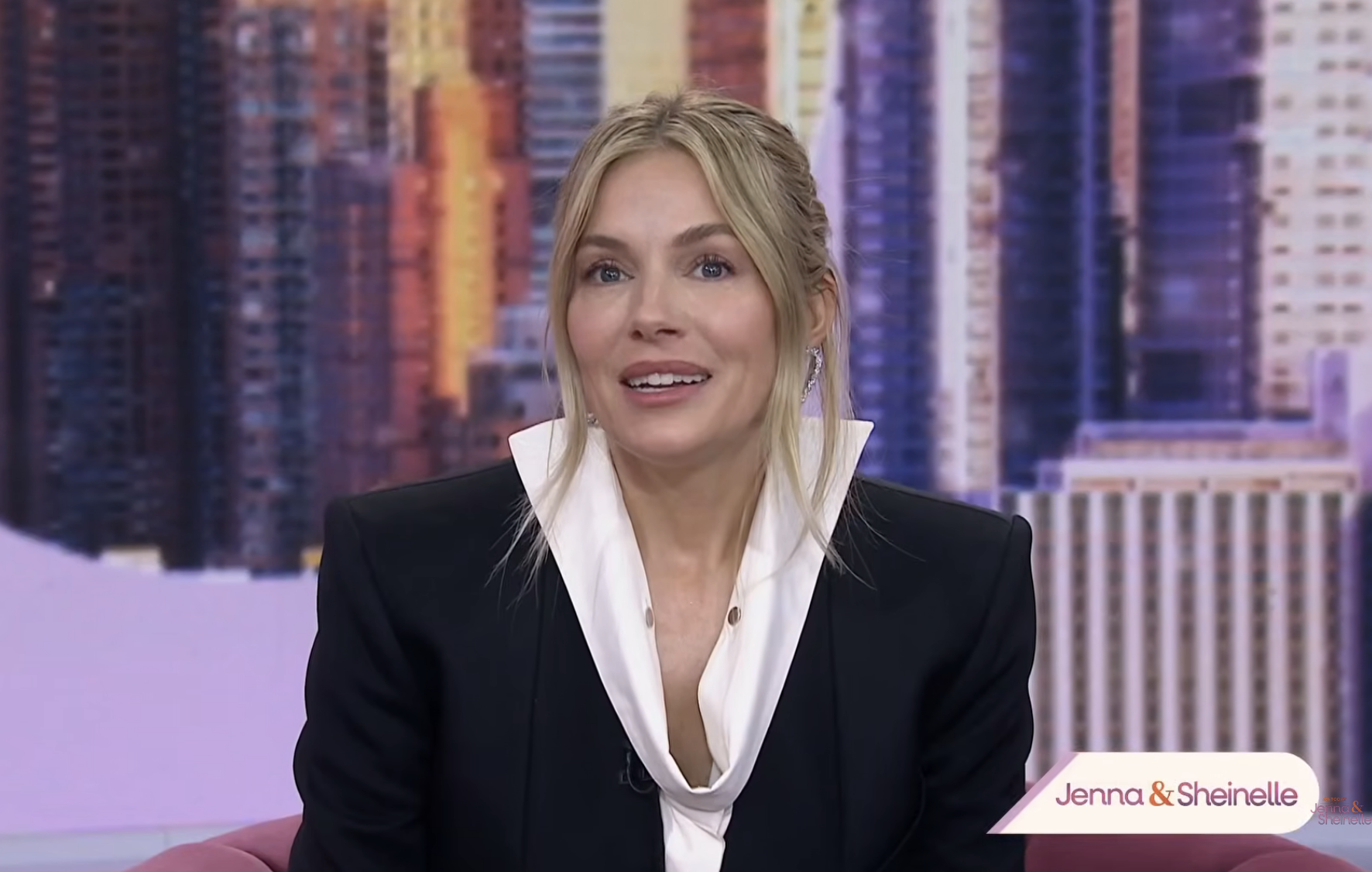 Person in a tailored black blazer with a white collar, seated on a set labeled "Jenna &amp;amp; Sheinelle," with a cityscape backdrop