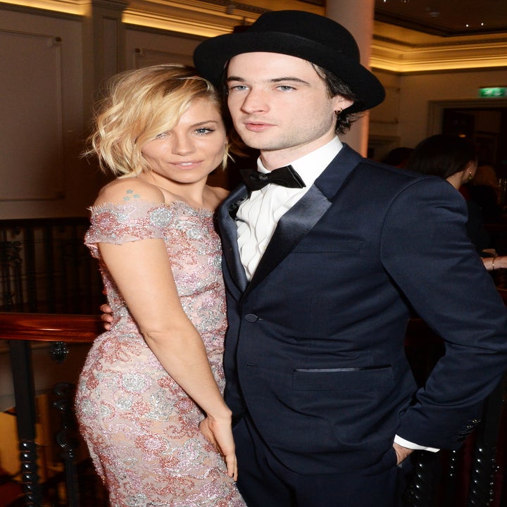 Two individuals posing together; the woman is wearing a floral-patterned, off-the-shoulder dress, and the man is in a suit with a bow tie and hat