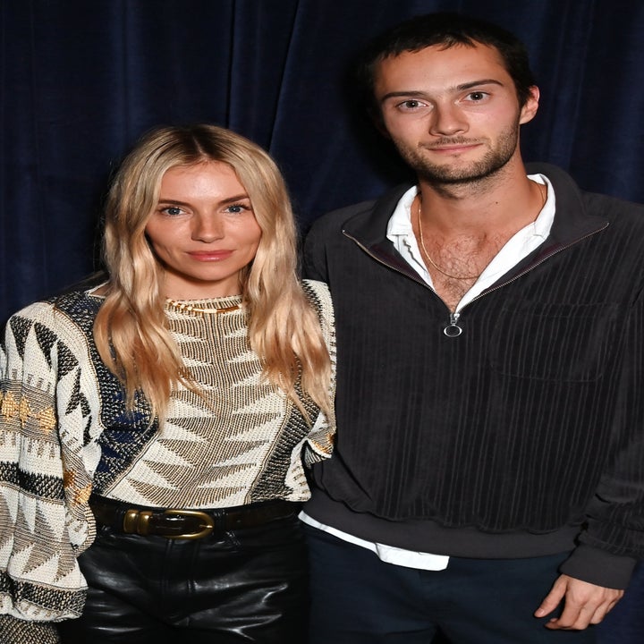 Two people standing together. The woman is wearing a patterned sweater and leather pants, while the man is dressed casually in a jacket and shirt