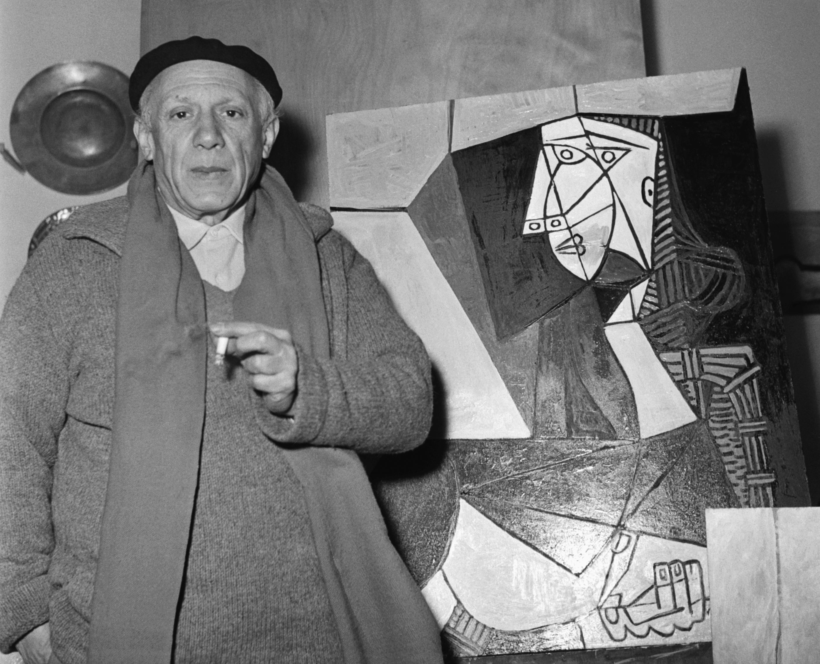 An older man stands beside a cubist painting, wearing a beret, scarf, and coat, holding a cigarette, likely in an art studio setting
