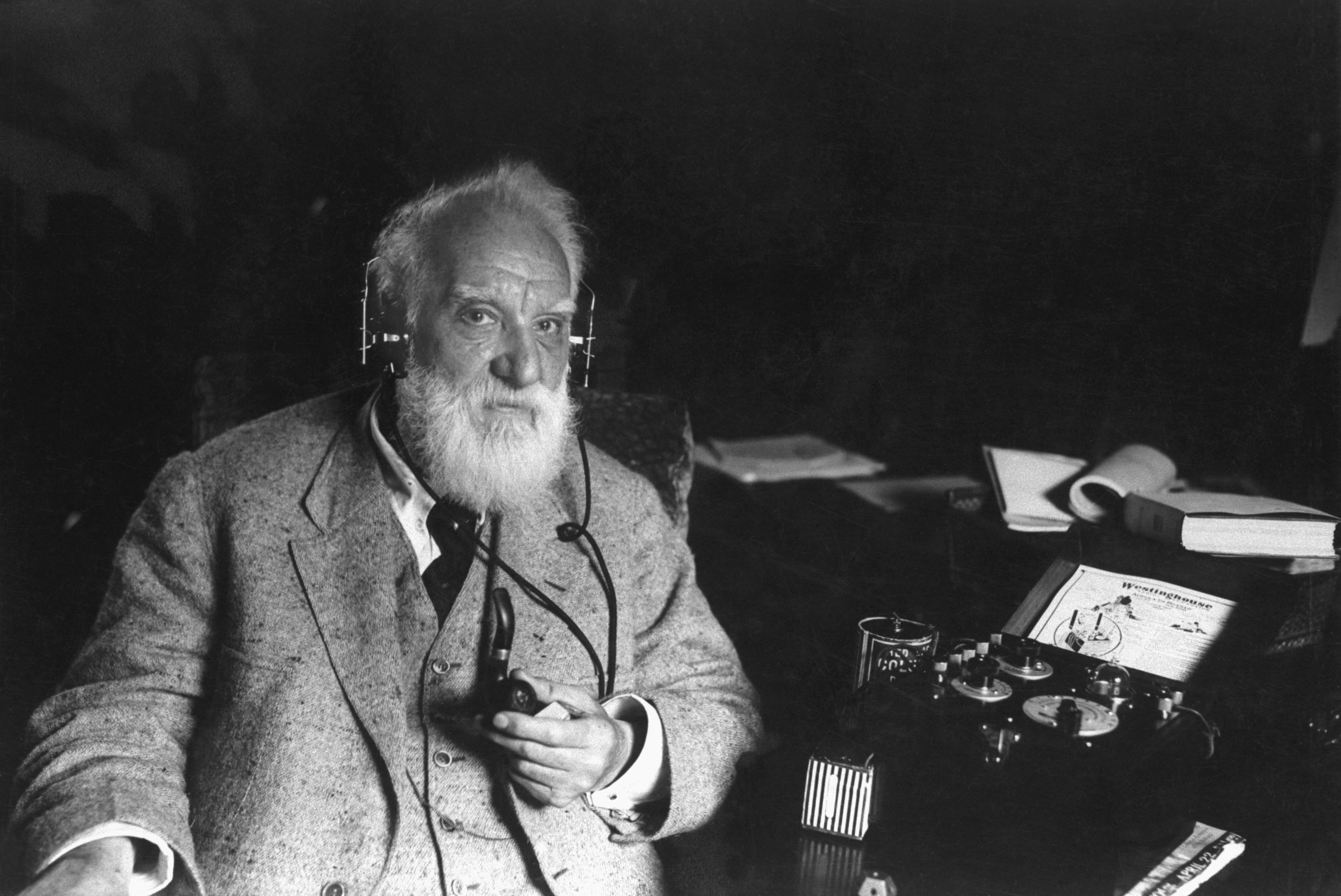 An older man with a beard wearing a suit and headphones, seated beside vintage communication equipment, holding a pipe