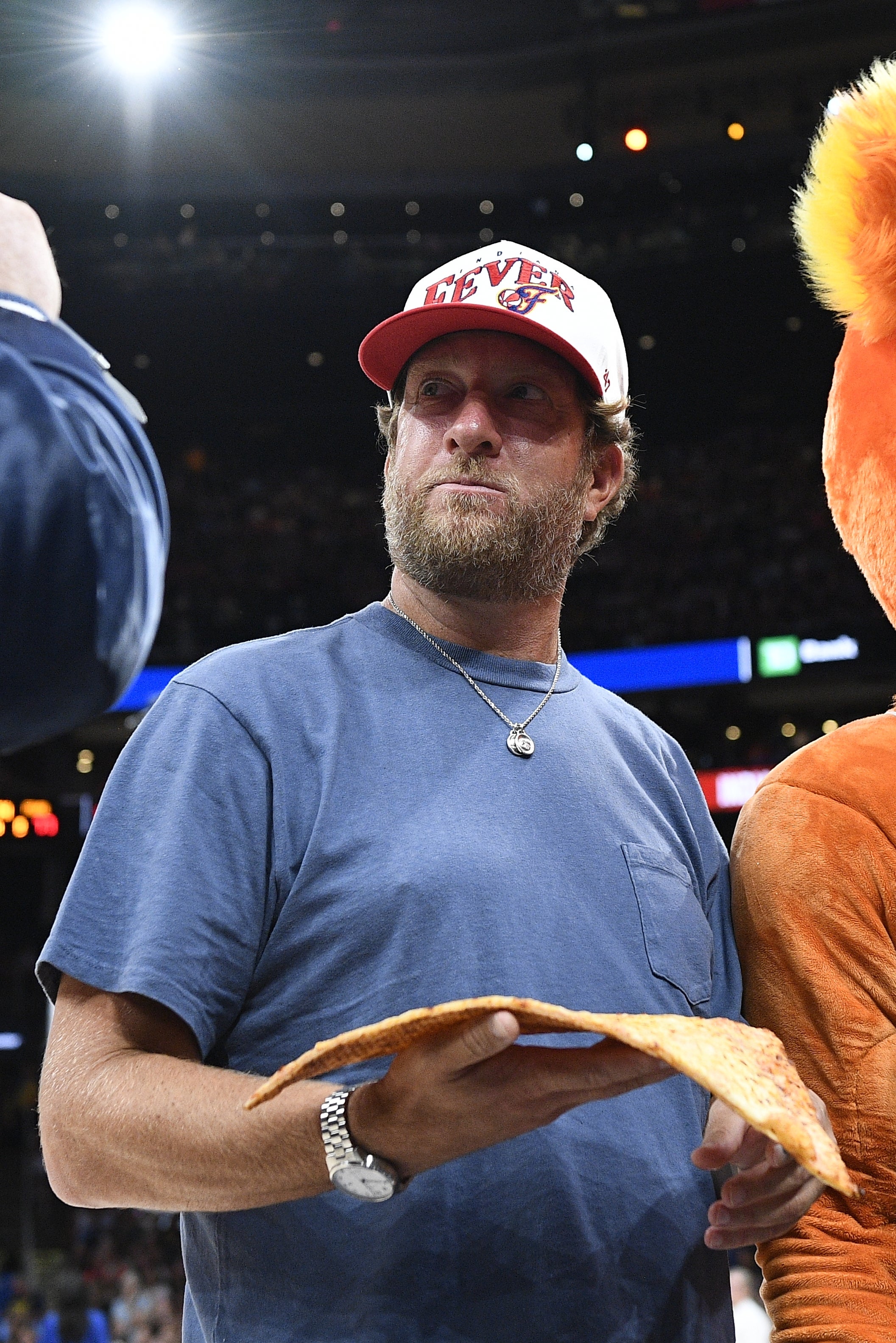 Person in casual attire holding a slice of pizza at a public event, wearing a cap and a necklace