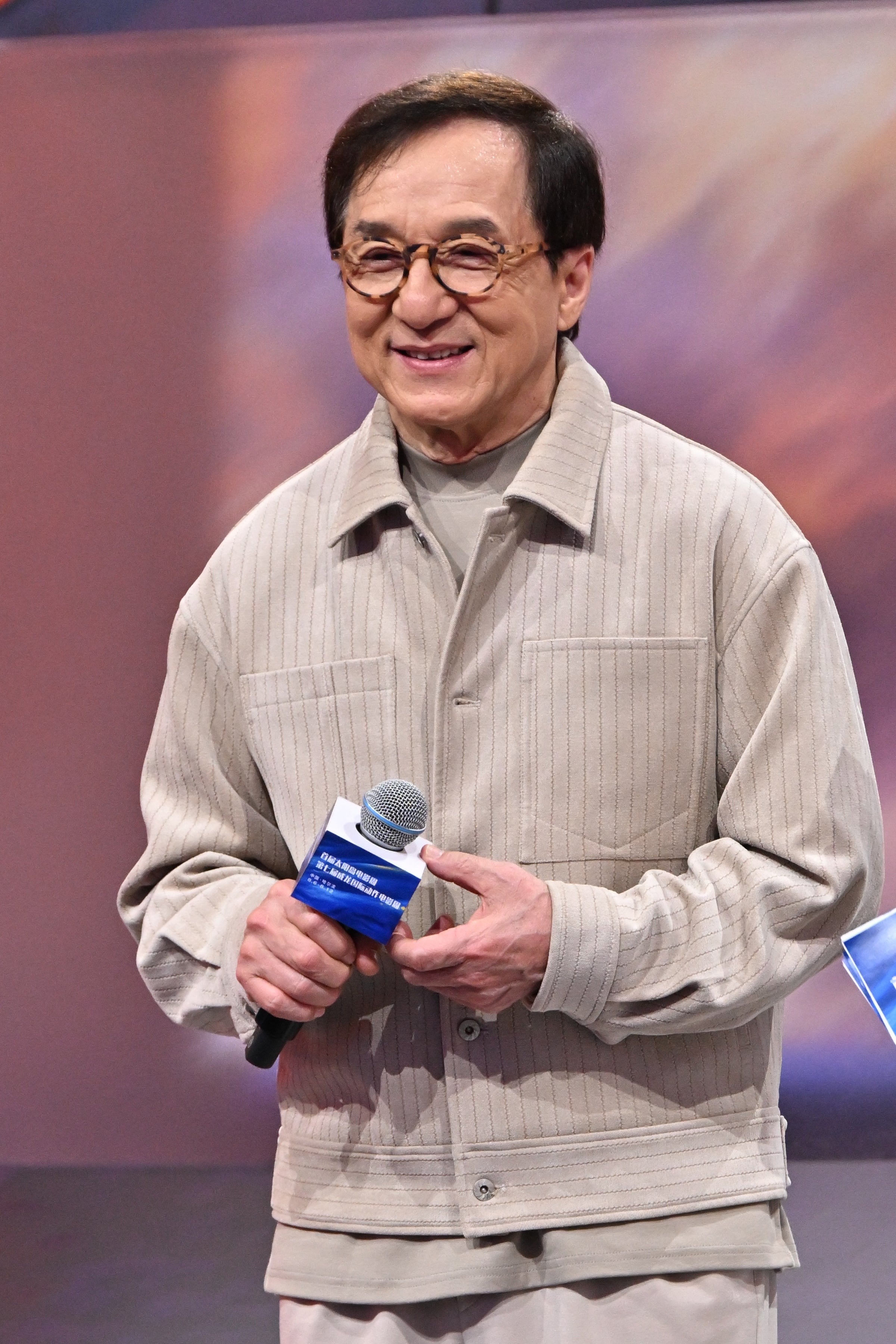A person smiling while holding a microphone, wearing a casual, ribbed jacket
