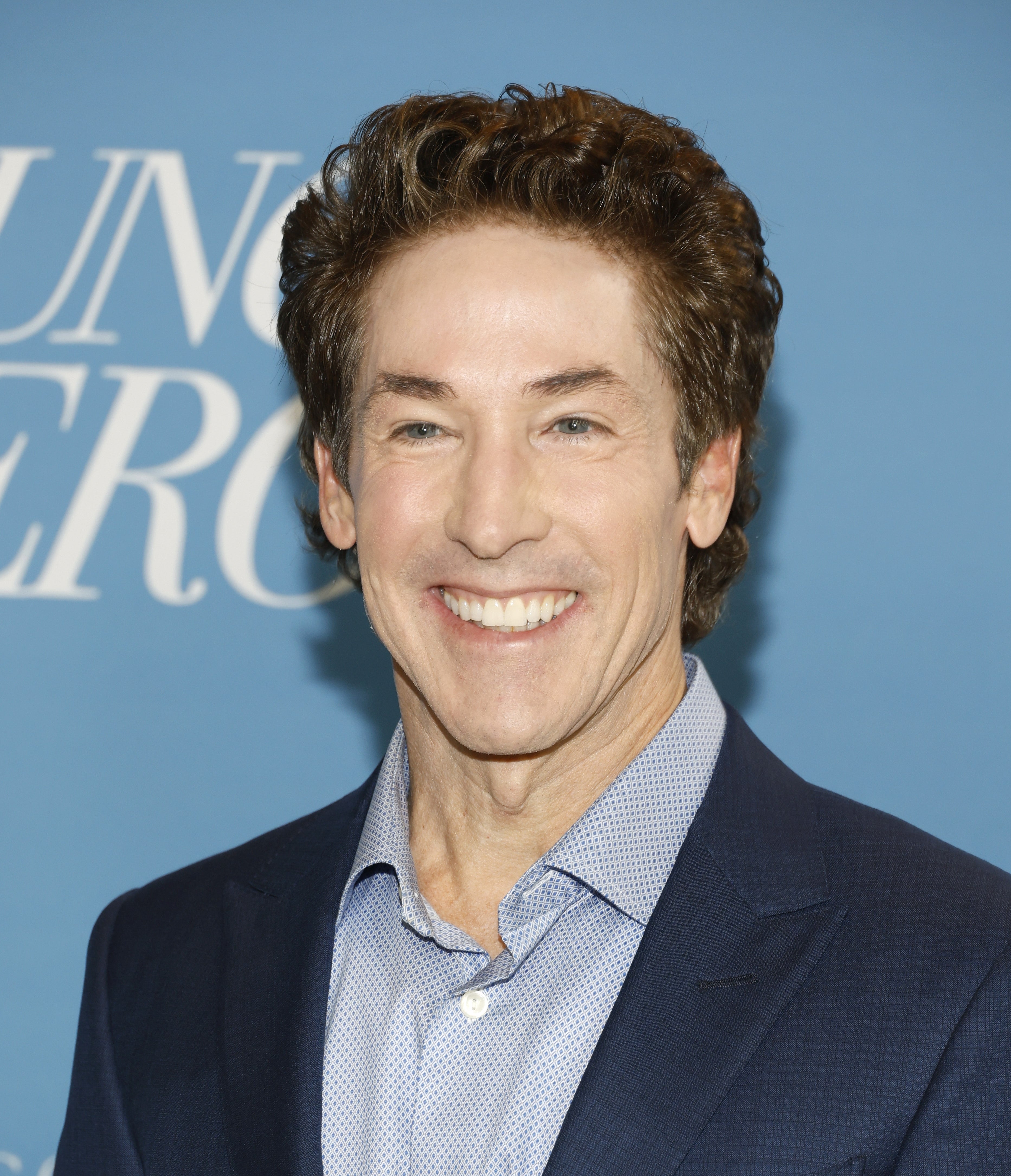 Person in a tailored suit smiling in front of a blue background with partial text