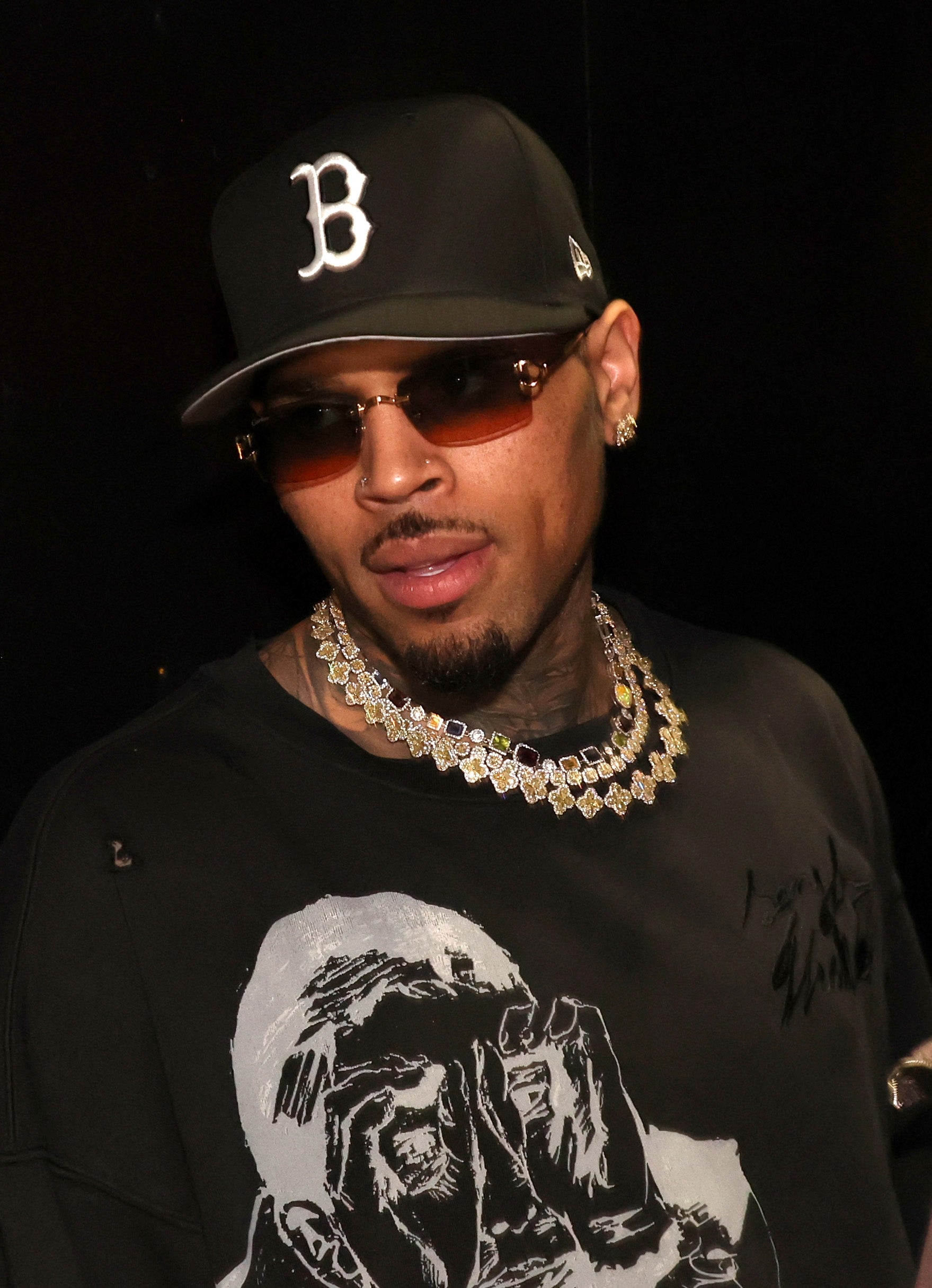 Man in casual black outfit with gold chains and sunglasses wearing a baseball cap, posing for the camera