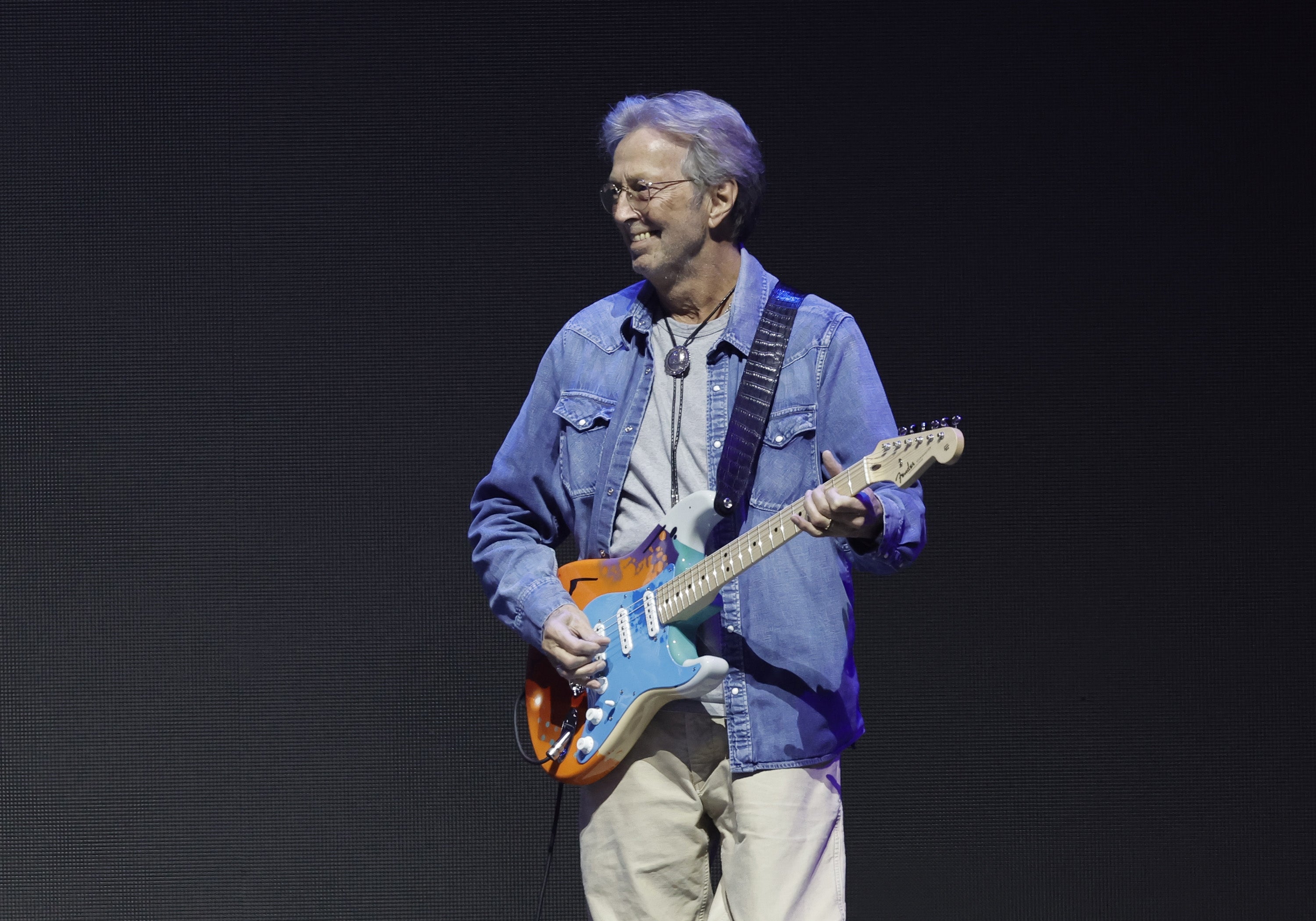 A musician in a casual denim jacket and pants plays an electric guitar on stage