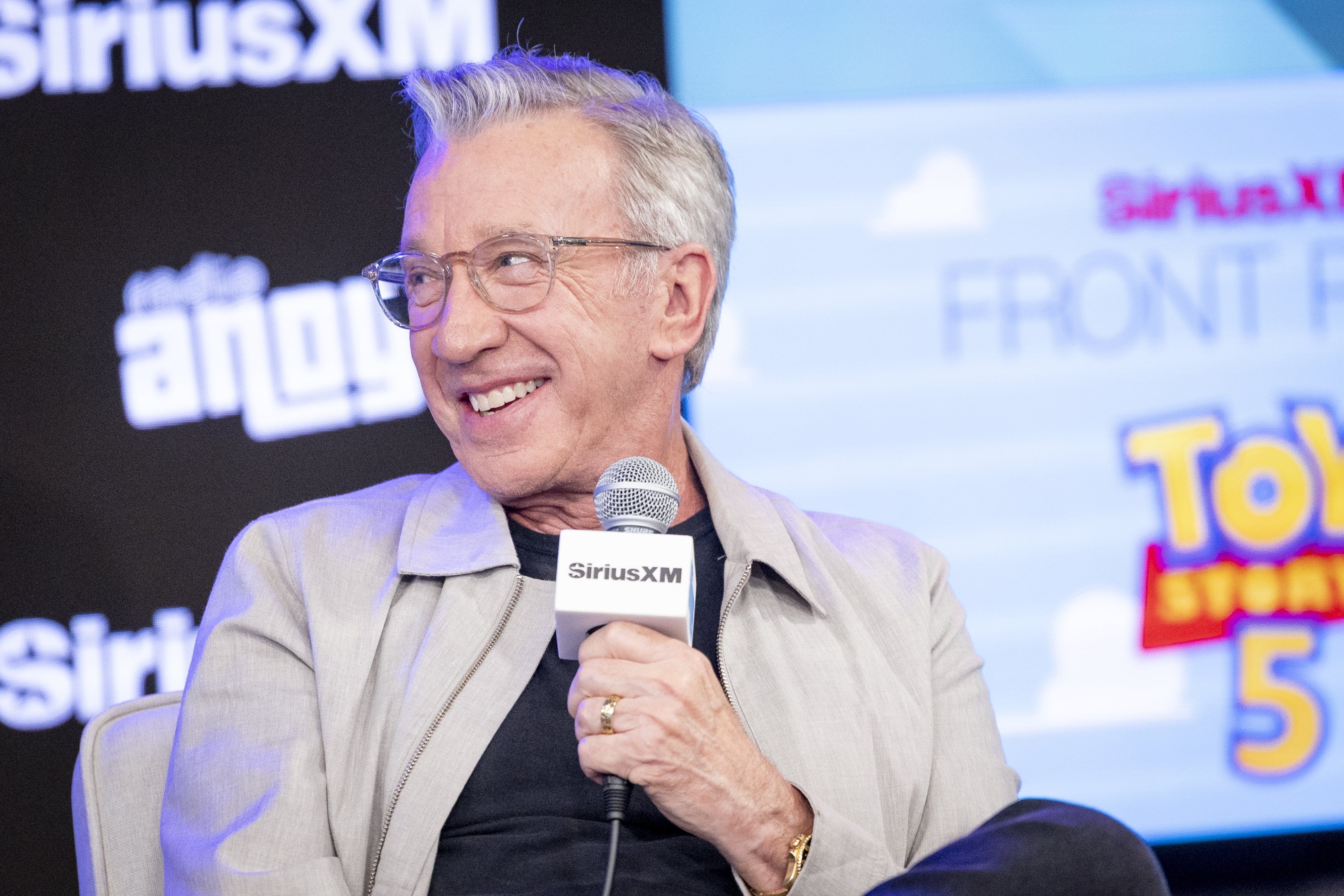 A person smiles while holding a SiriusXM microphone at a "Toy Story 5" event. They're wearing a light jacket and glasses