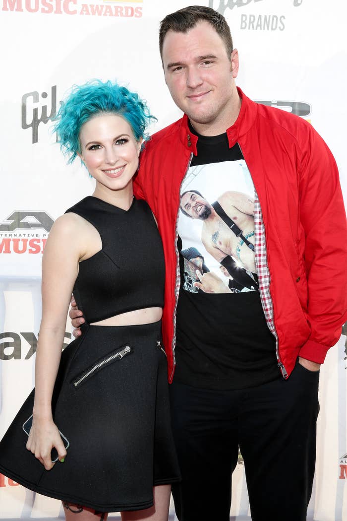 A person with short, vibrant blue hair in a black crop top and skirt poses with another person in a red jacket and graphic tee on a red carpet