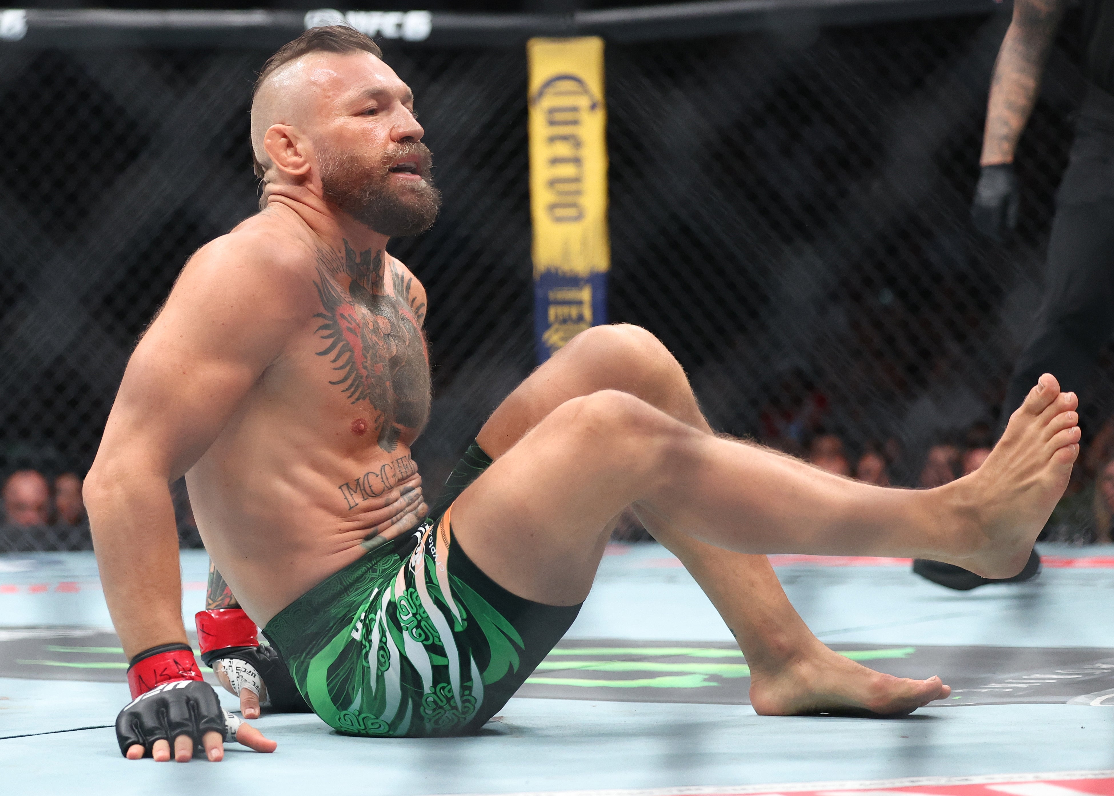 Athlete sitting in an octagon during a UFC match, wearing green patterned shorts and gloves, with multiple tattoos visible