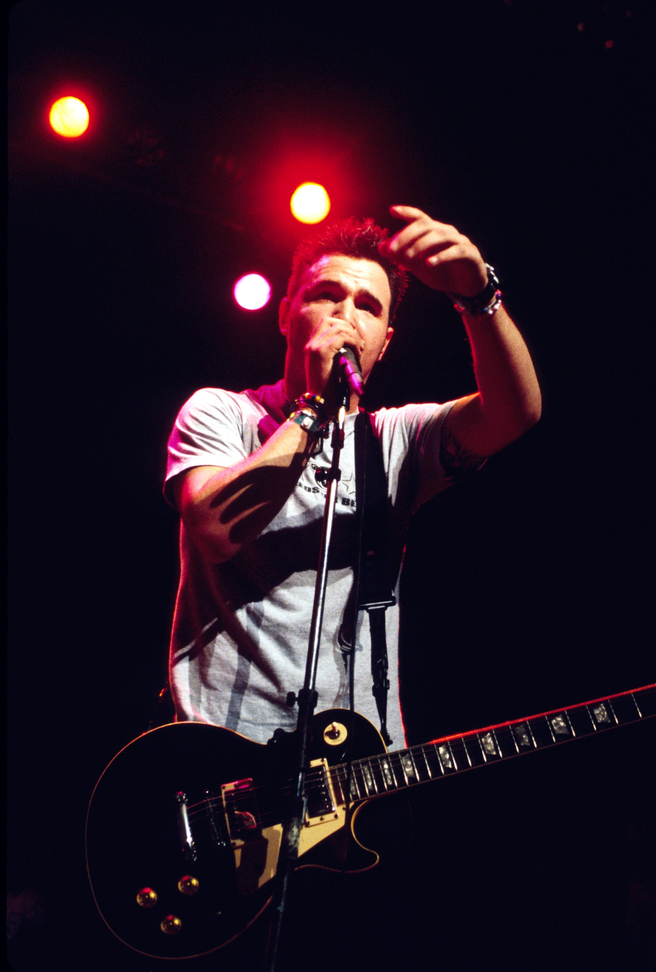Musician performing on stage with a guitar, gesturing to the audience, wearing a casual t-shirt