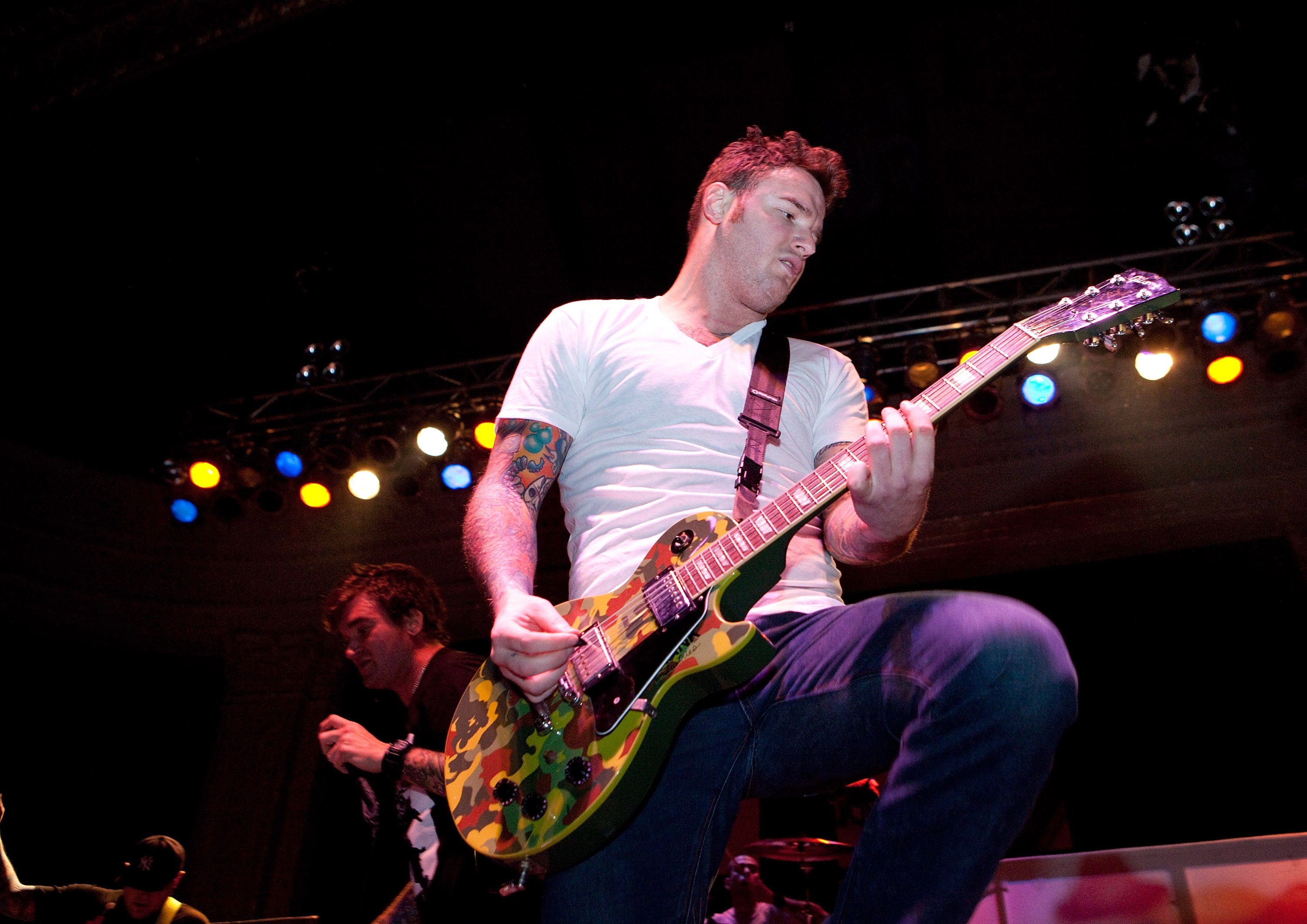 Guitarist passionately playing on stage, wearing a white T-shirt and jeans, with colorful stage lighting in the background