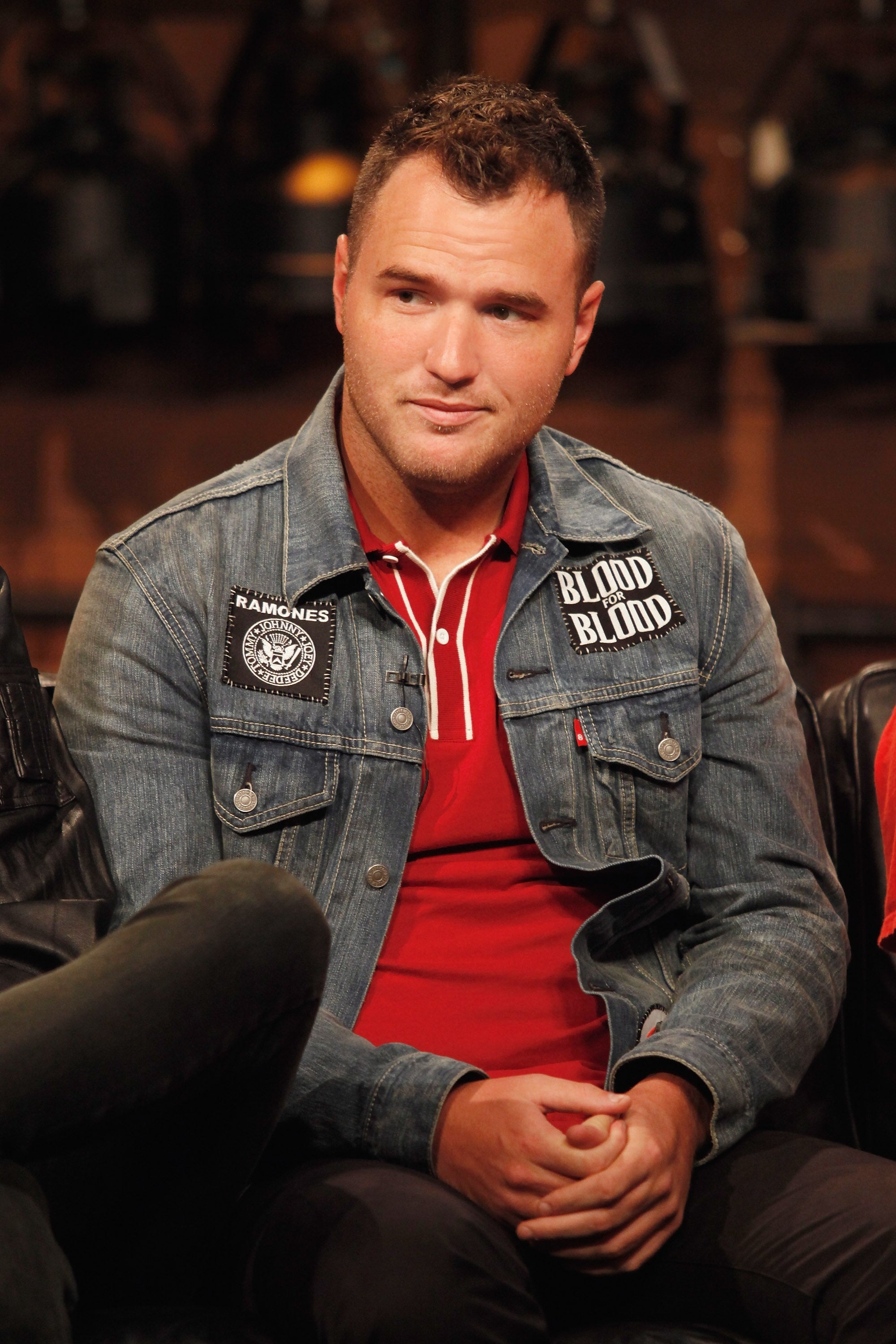 Person seated wearing a denim jacket with patches and a red shirt underneath, in a casual setting