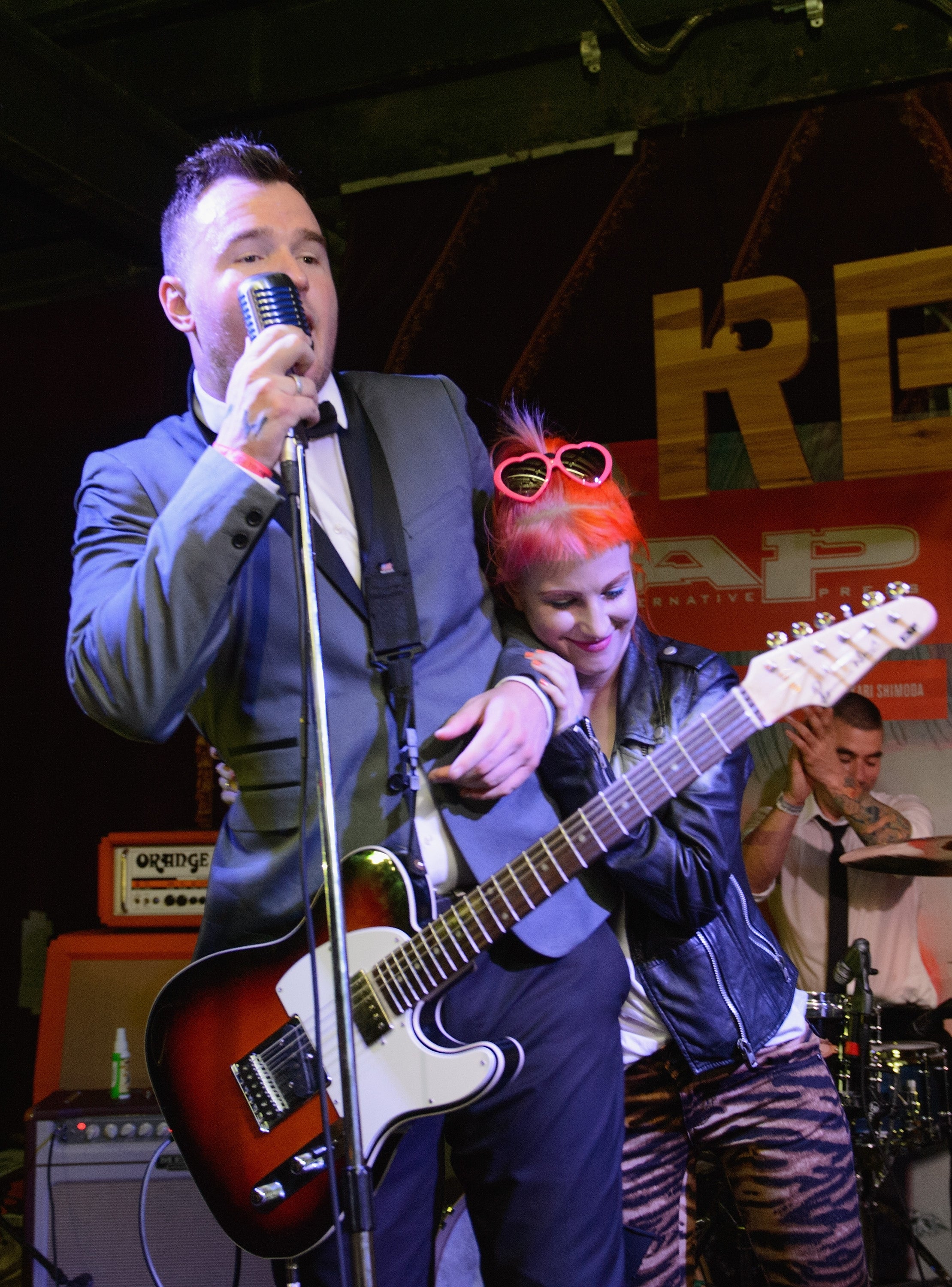 A musician in a suit sings into a vintage microphone, while a woman with colorful hair and funky eyewear leans on him, both enjoying a live performance