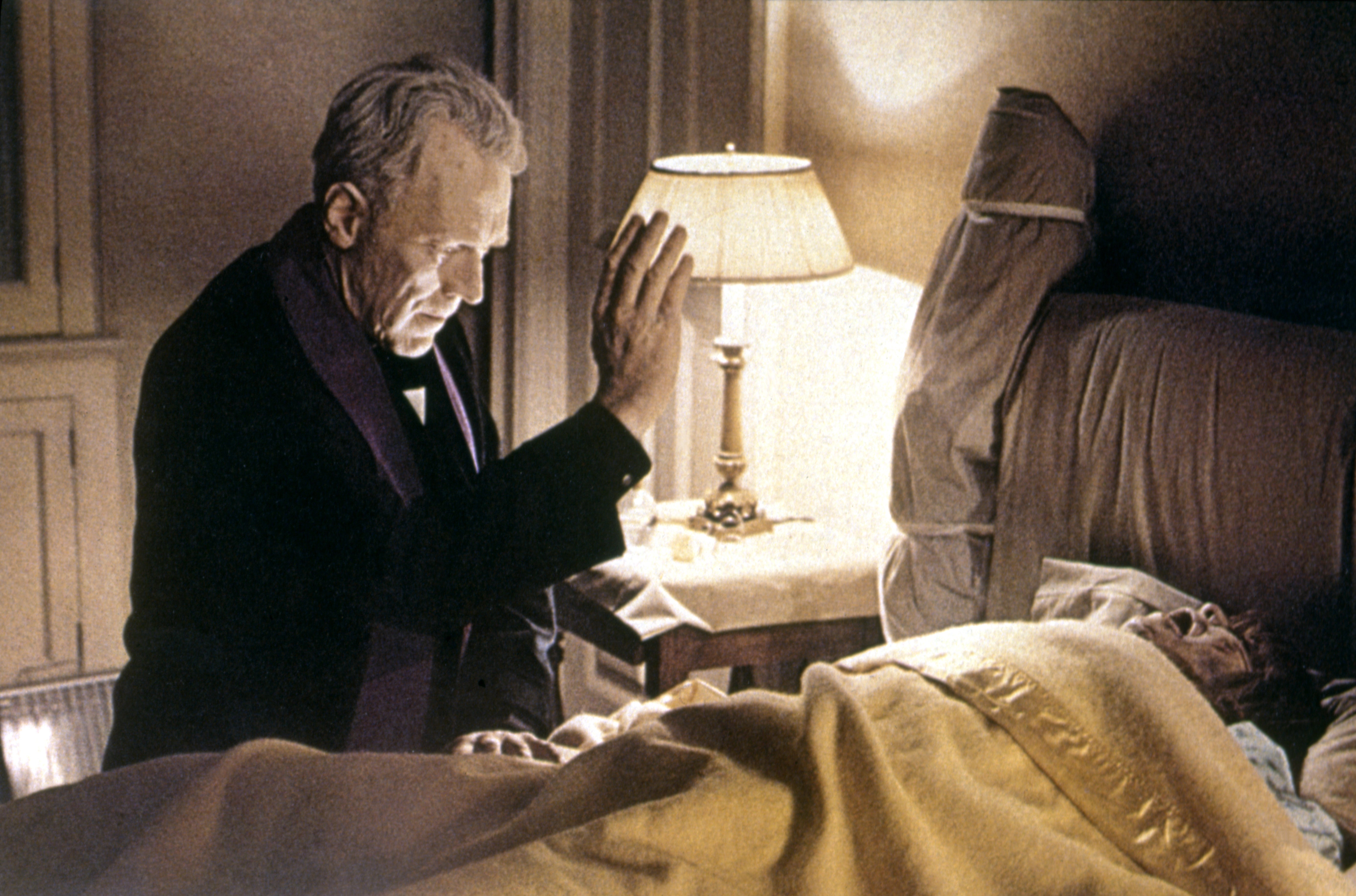 Father Lankester Merrin stands by a bed, holding his manus up implicit Regan MacNeil, lying nether the covers successful a country from the movie 'The Exorcist,' 1973