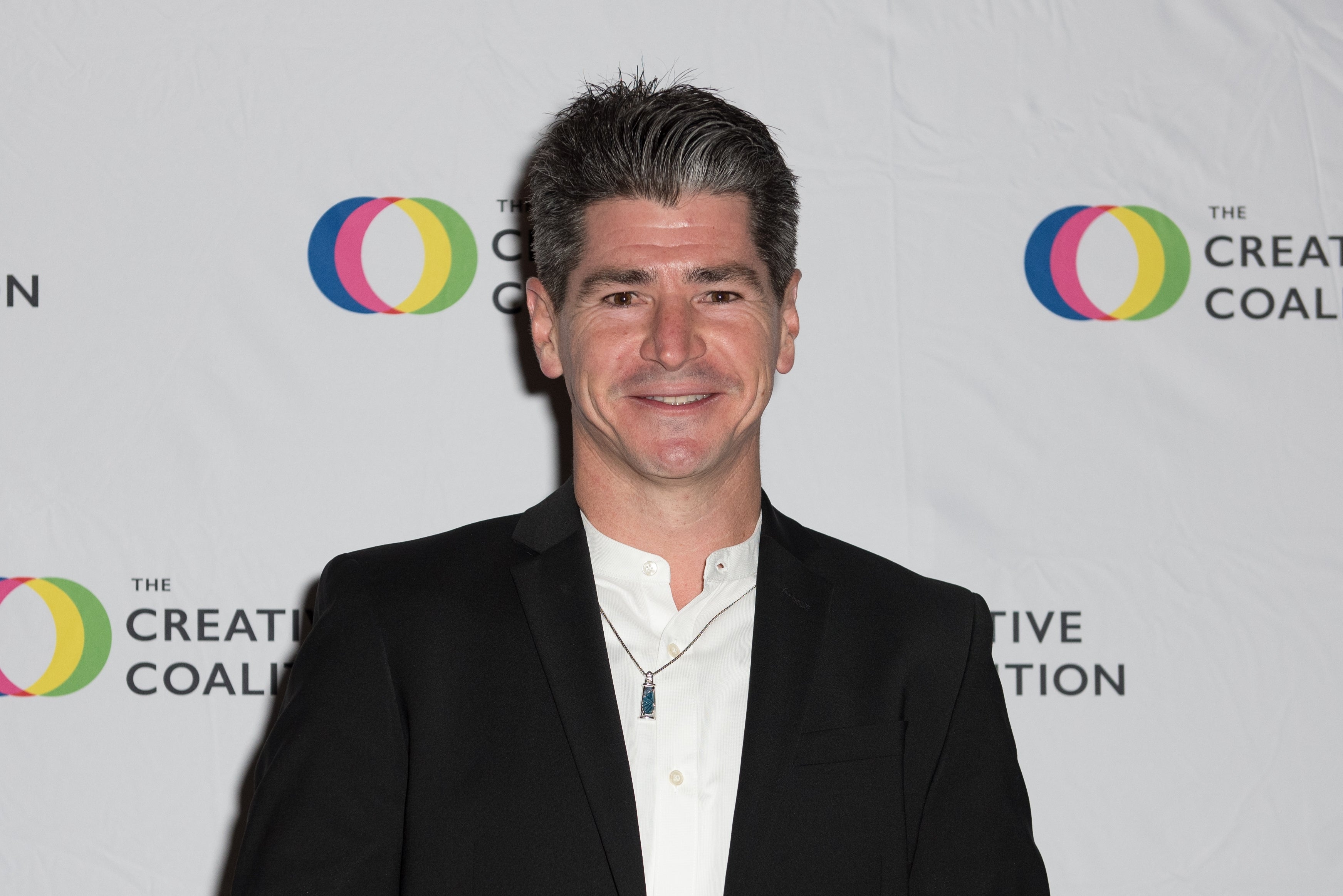 Man in a suit with a necklace, standing in front of a backdrop with "The Creative Coalition" logo