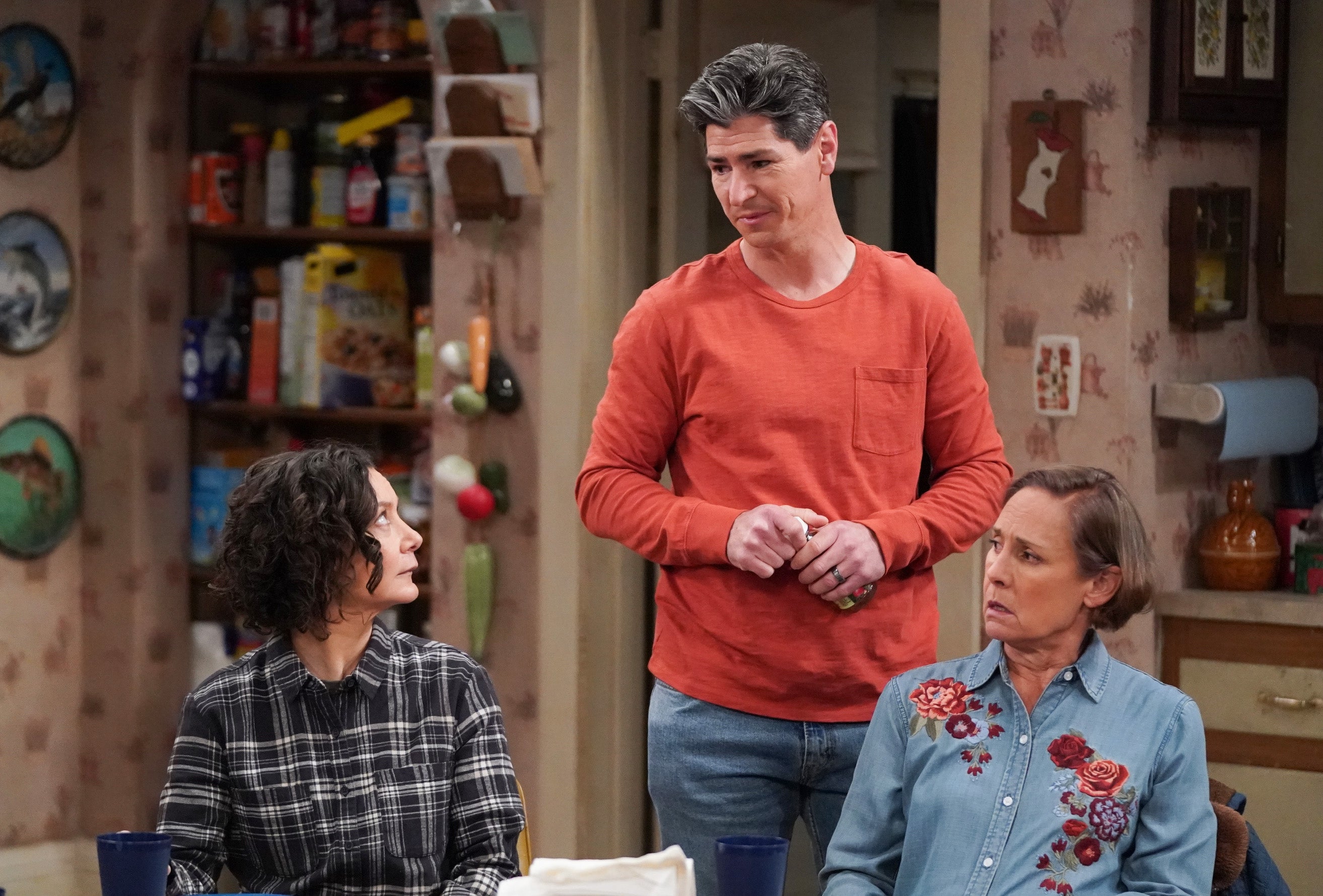 Three people are in a kitchen, having a serious conversation. One person is standing, while the others are sitting, listening attentively