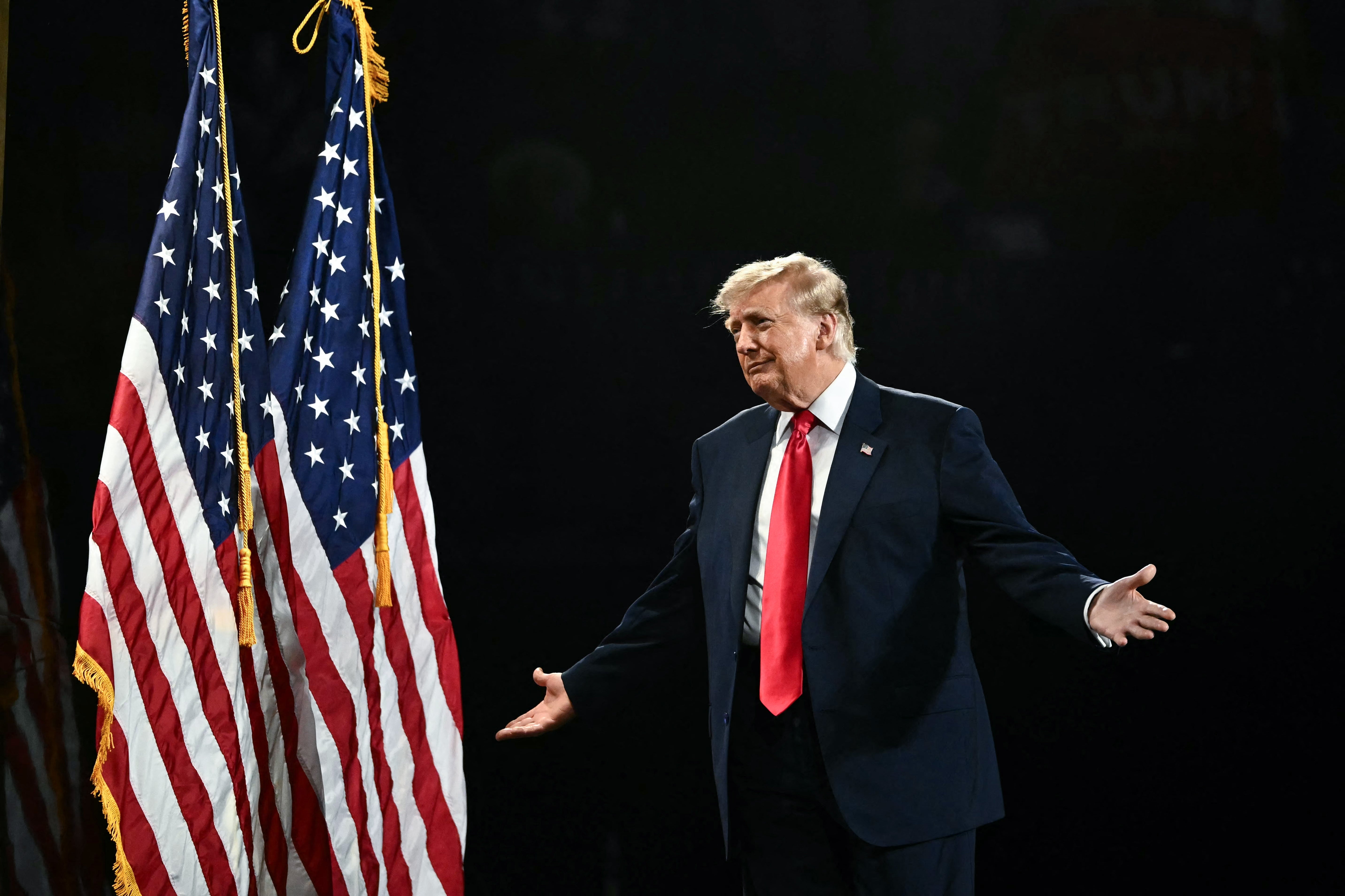 En mann i dress med rødt slips står på scenen med amerikanske flagg i bakgrunnen og gestikulerer med åpne armer