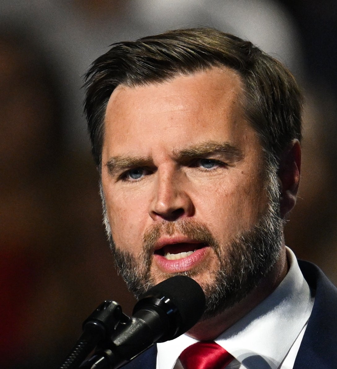JD Vance i dress snakker lidenskapelig ved en mikrofon under et arrangement, og holder opp en finger for å understreke