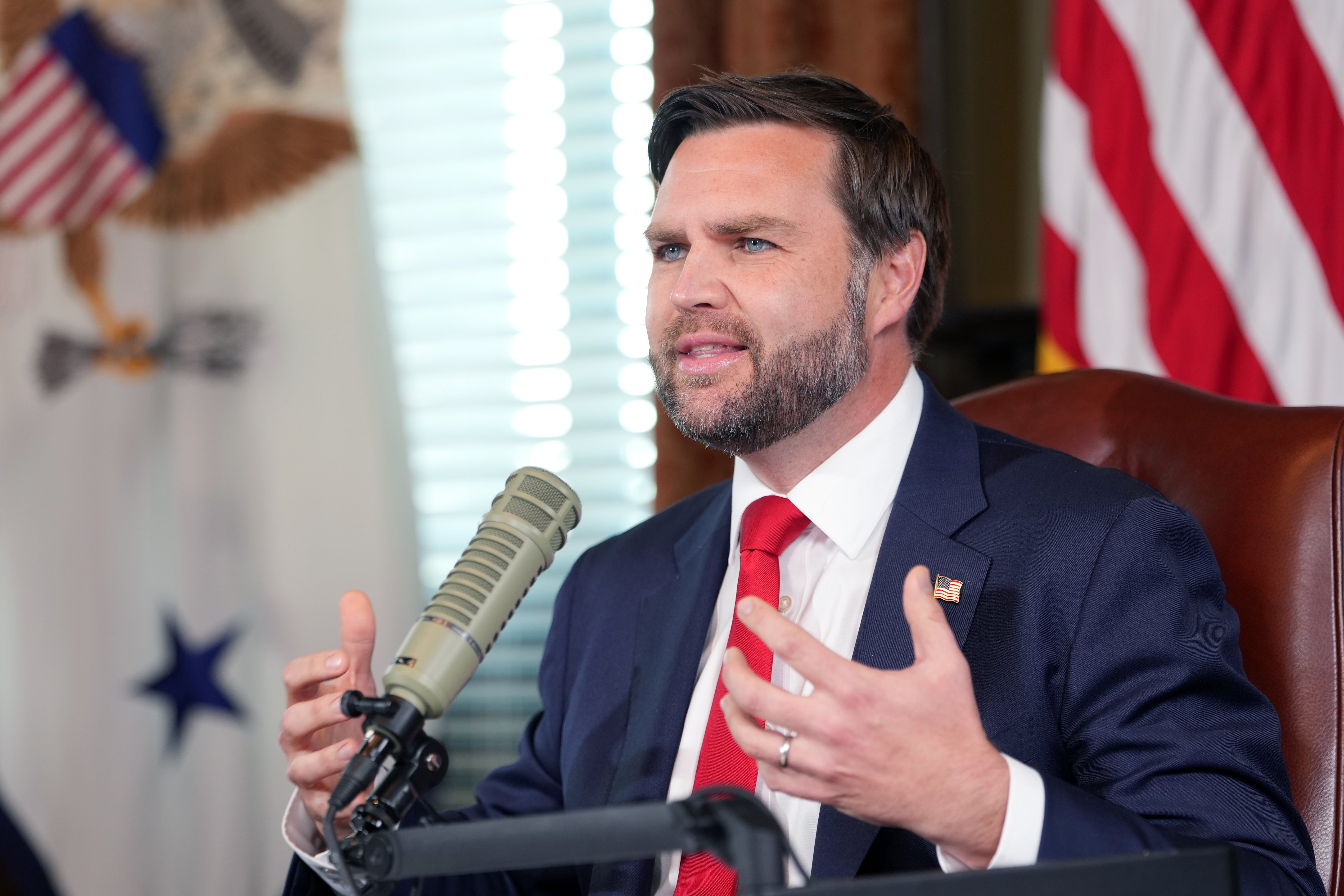 JD Vance i dress og slips snakker lidenskapelig inn i en mikrofon, gestikulerer med hendene, med flagg synlige i bakgrunnen