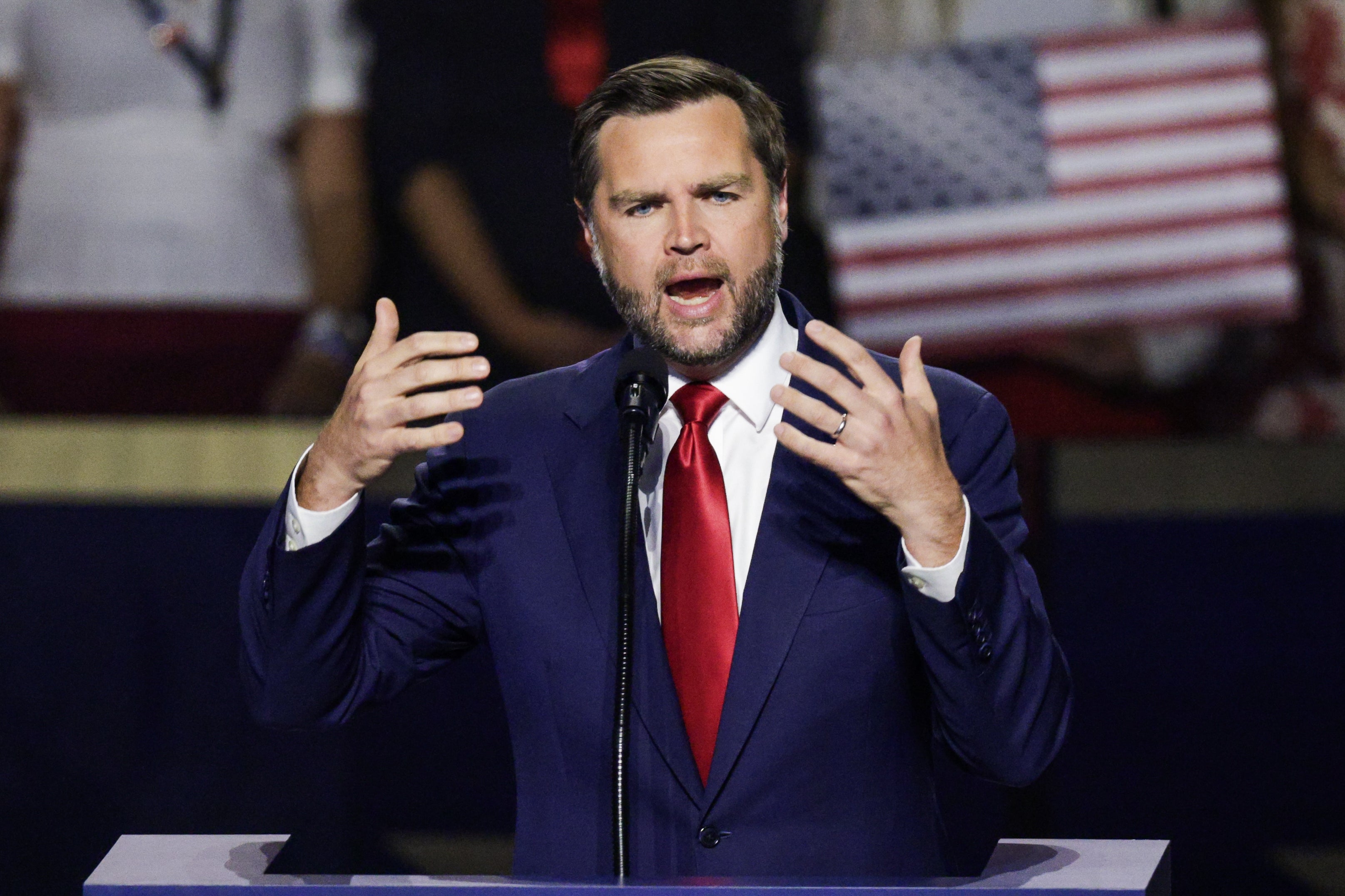 JD Vance i dress og rødt slips snakker ved et podium og gestikulerer med hendene. Et amerikansk flagg er synlig i bakgrunnen