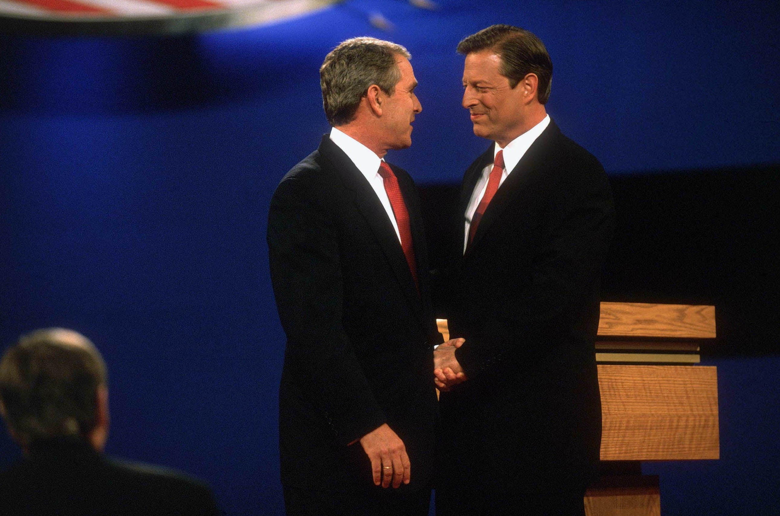 George W. Bush og Al Gore i dress og slips håndhilser på scenen under en debatt. En person ser på fra forgrunnen