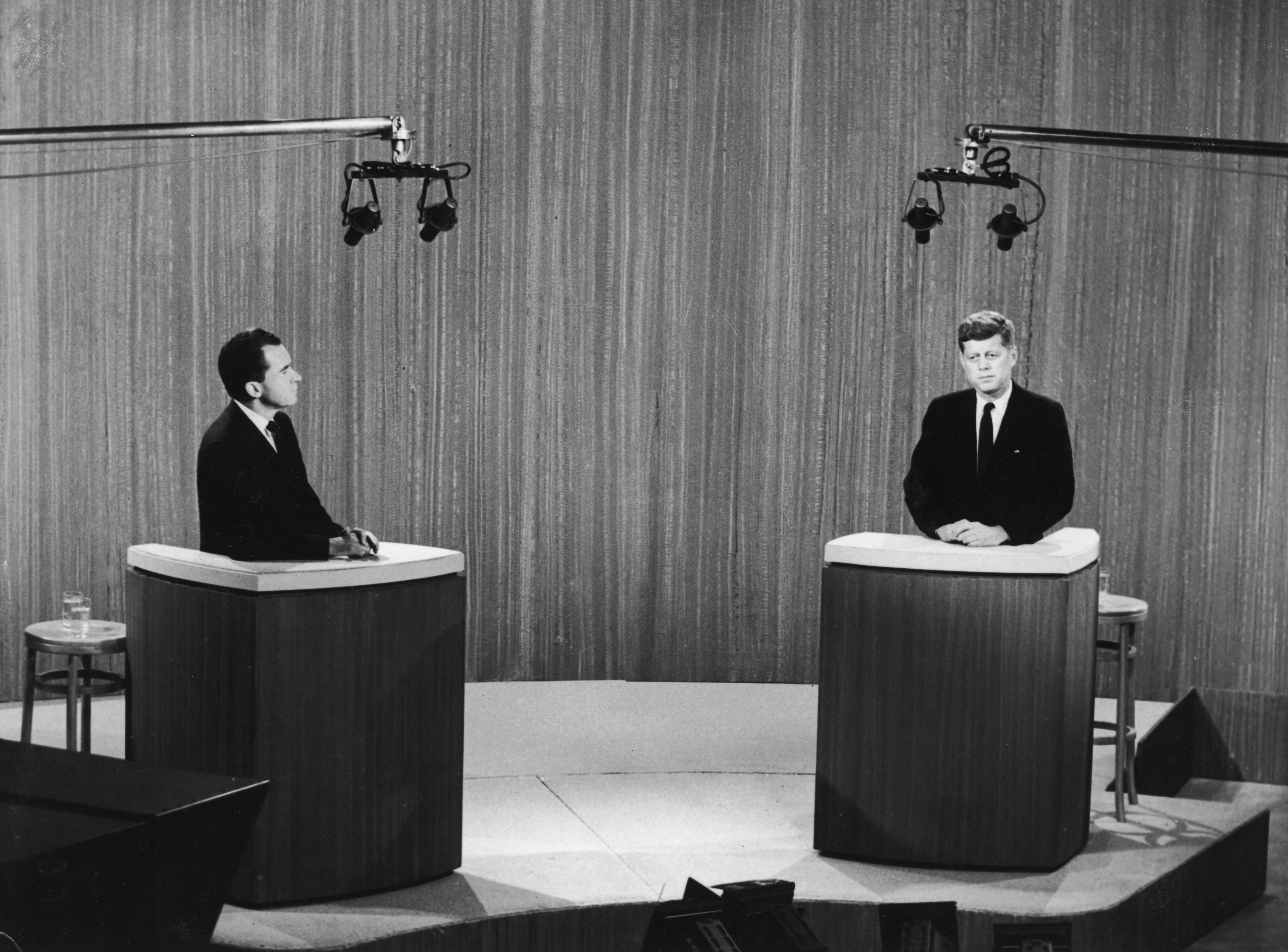 Nixon og Kennedy, i dress, står bak podier under en TV-debatt på settet, med overlys synlig