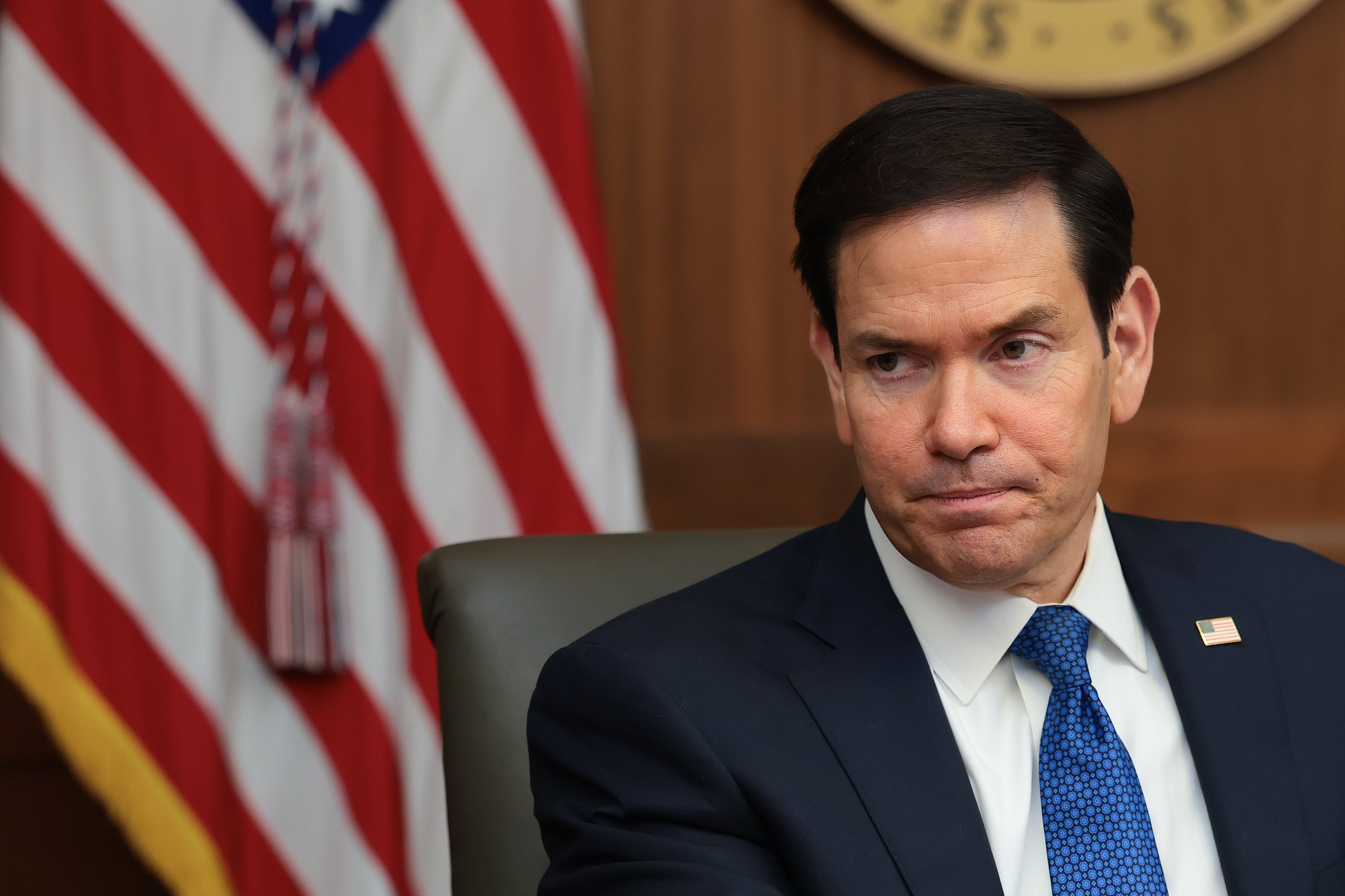 Marco Rubio i en formell dress sitter med et amerikansk flagg synlig i bakgrunnen, og virker gjennomtenkt eller bekymret