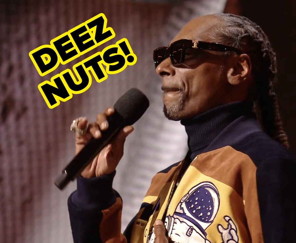 Person wearing sunglasses and a turtleneck with a colorful jacket, holding a microphone and speaking, likely at an event or performance