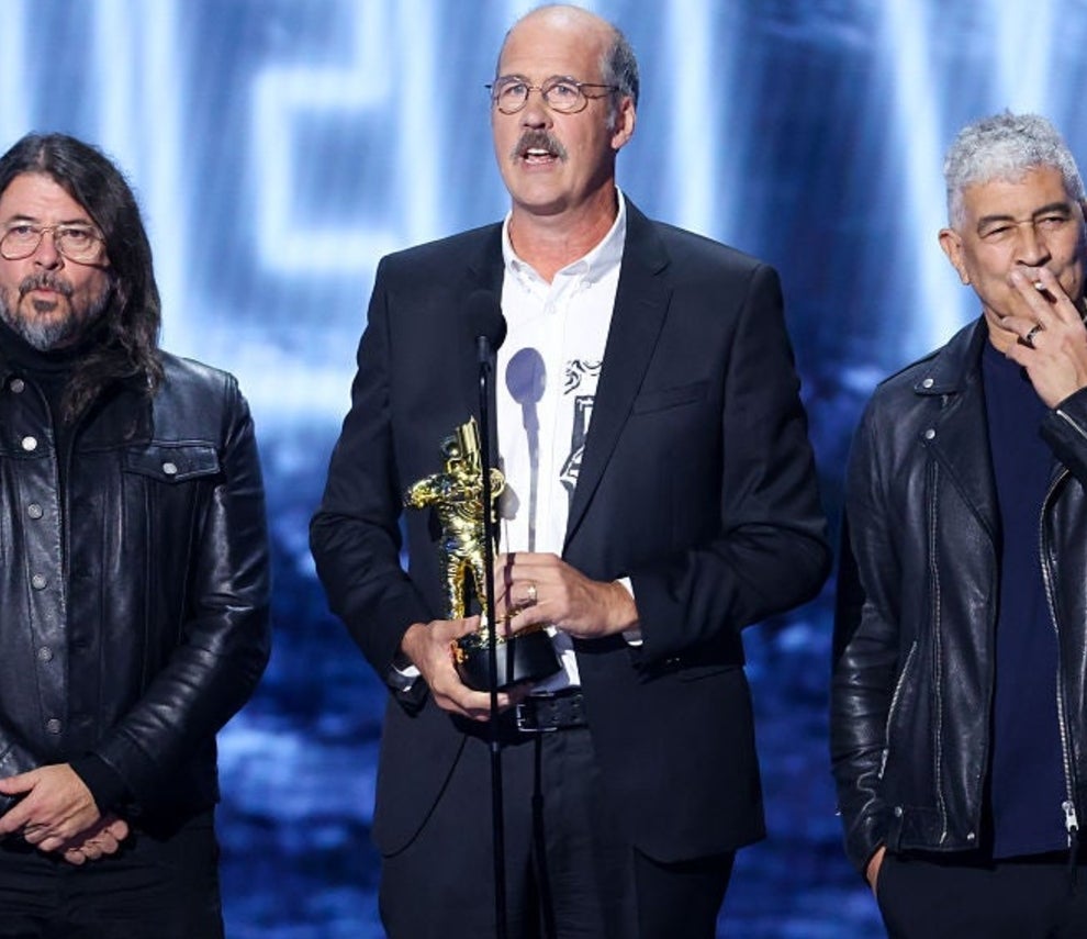 Three men stand on stage; one holds an award. They wear black leather jackets and speak into a microphone at a public event