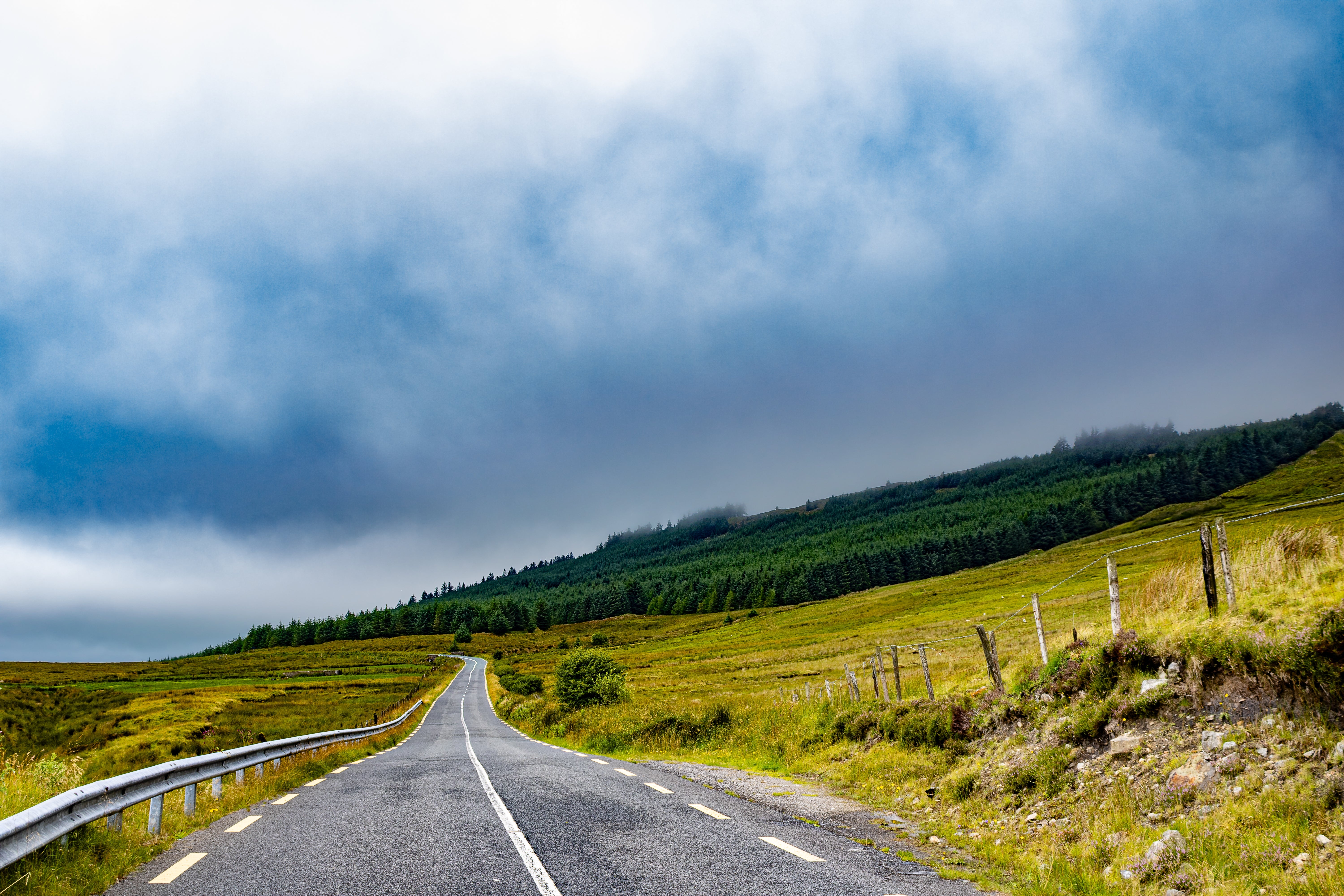 Scenic rural road stretching through green hills under a wide, cloudy sky, offering a tranquil travel view