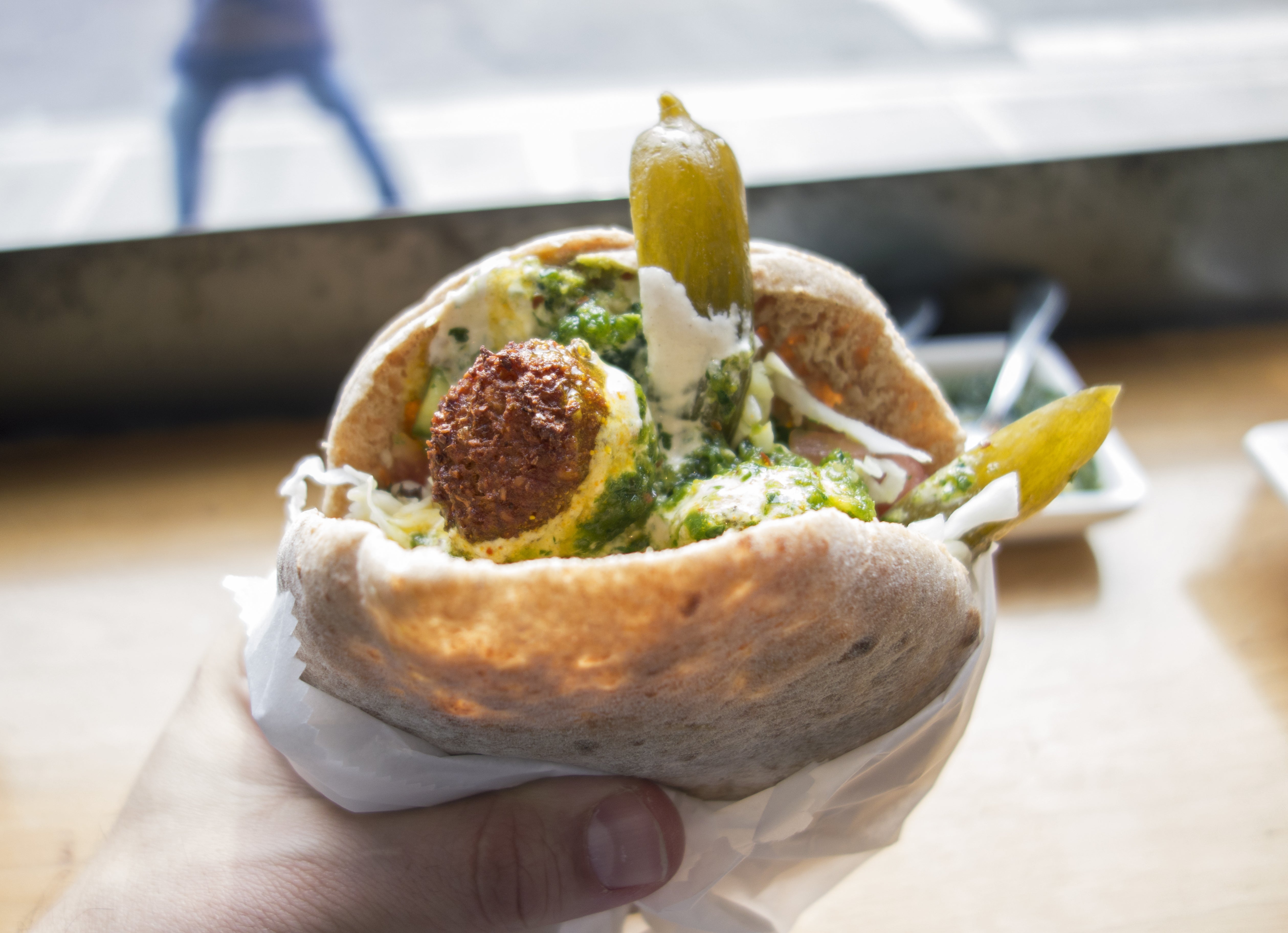 Pita filled with falafel, pickles, and vibrant green sauce, held over a wooden counter near a window with a blurred street view