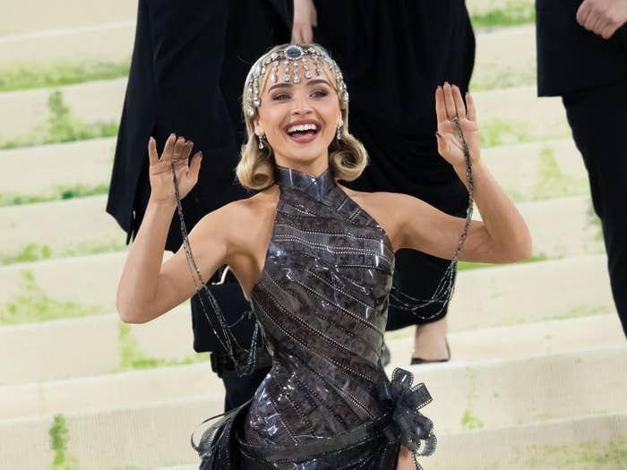 Person on red carpet in metallic, futuristic dress with headpiece, posing playfully with hands raised