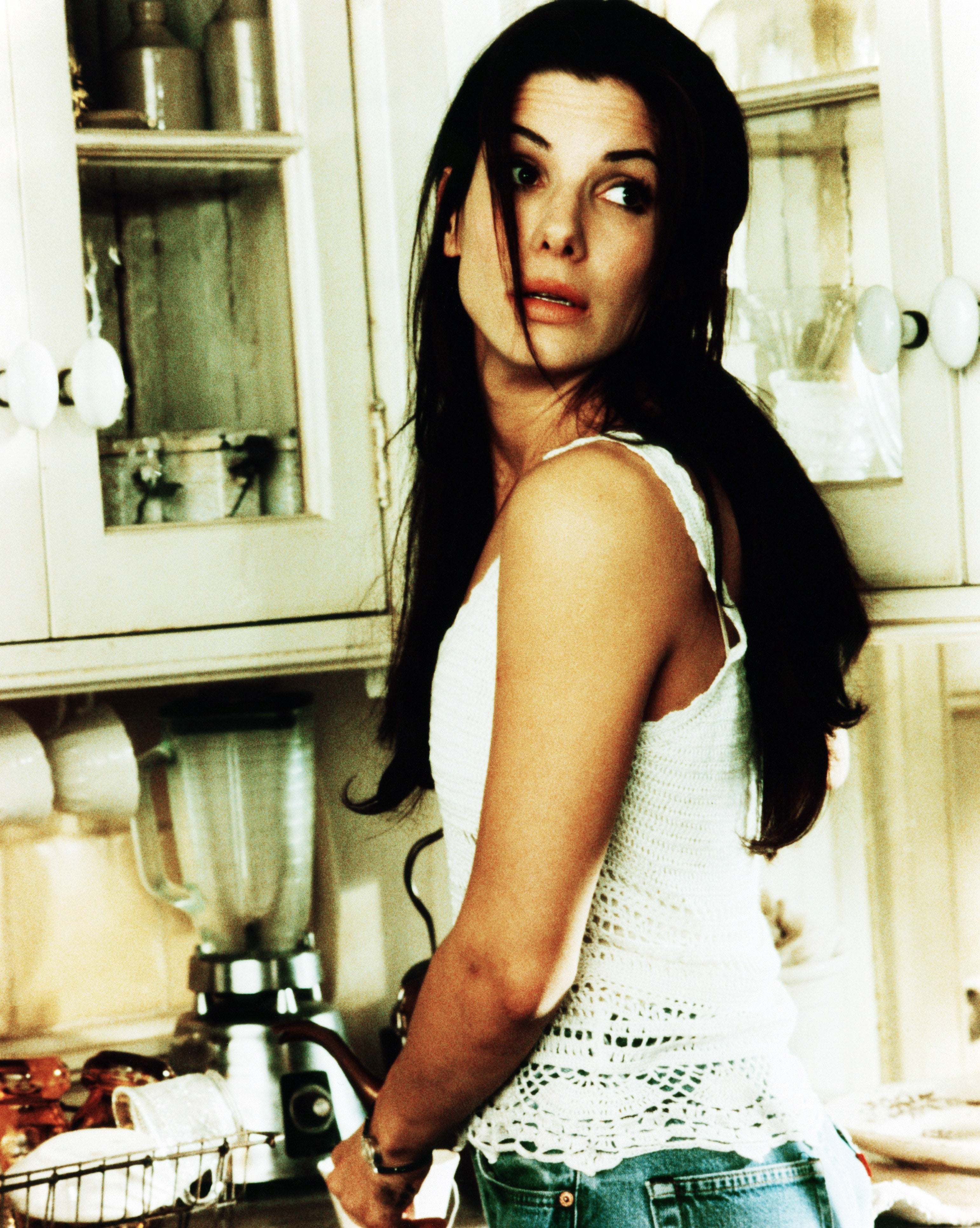 Person with long hair in a kitchen, wearing a sleeveless crocheted top and jeans, looking off to the side. A blender and kitchen items are in the background