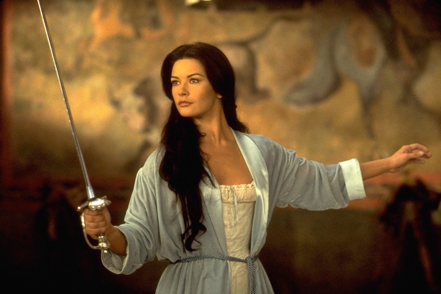 A person holding a sword in a poised stance, wearing a flowing robe, set against a mural background