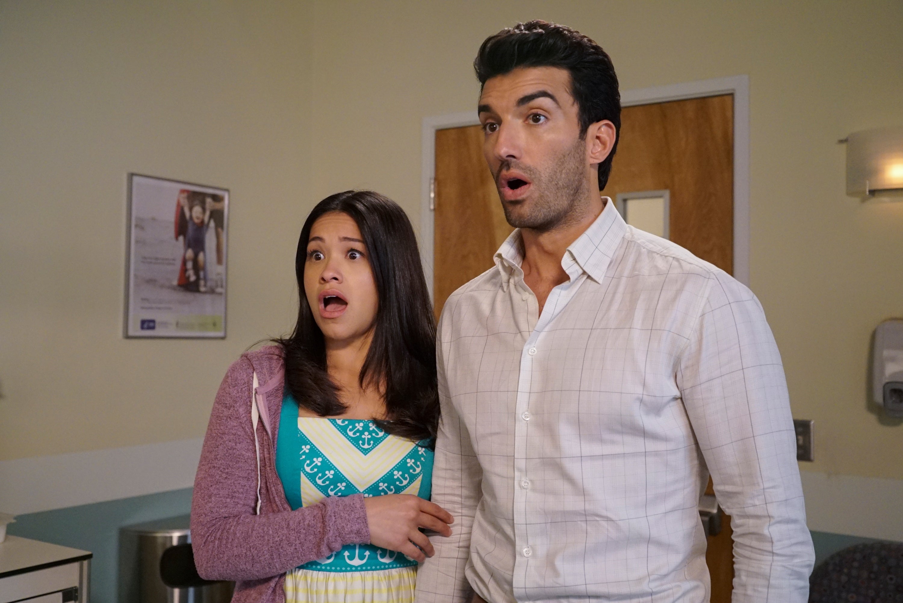 Two actors in casual attire, looking surprised, stand in a room with a poster and door in the background