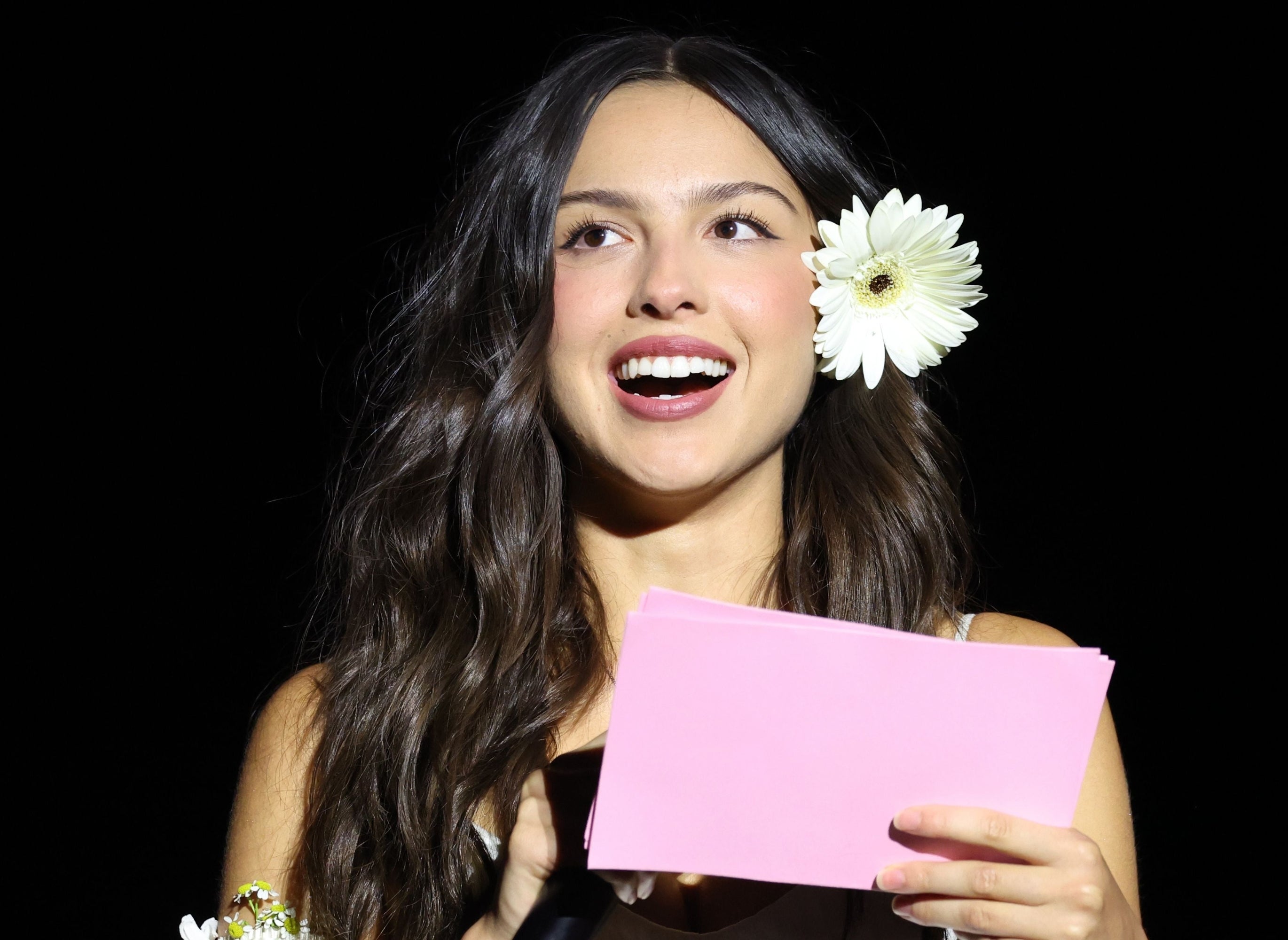 Person holding a microphone and a pink card, with a daisy in their hair, speaking on stage