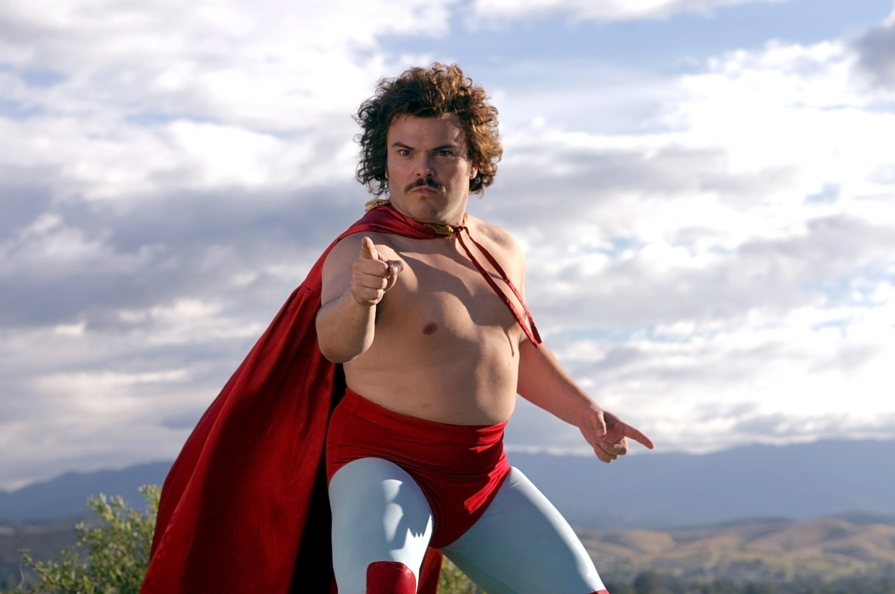A man poses confidently in a superhero costume with a red cape and tights, pointing forward against a scenic outdoor backdrop