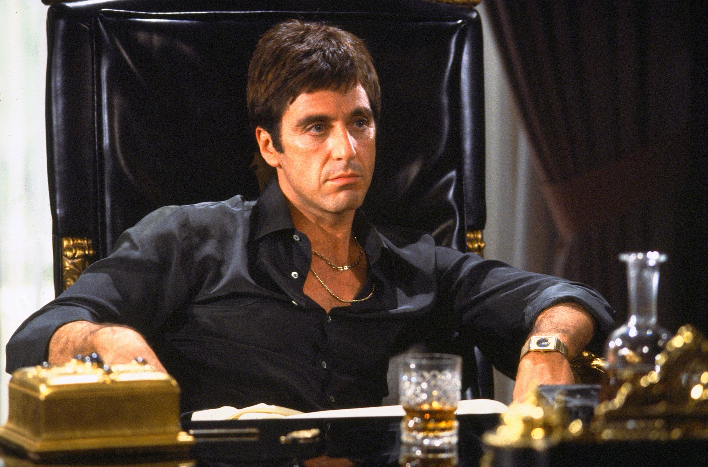 Man sitting at a desk, wearing a black shirt with an intense expression, surrounded by luxurious items like a gold telephone and a glass of liquor