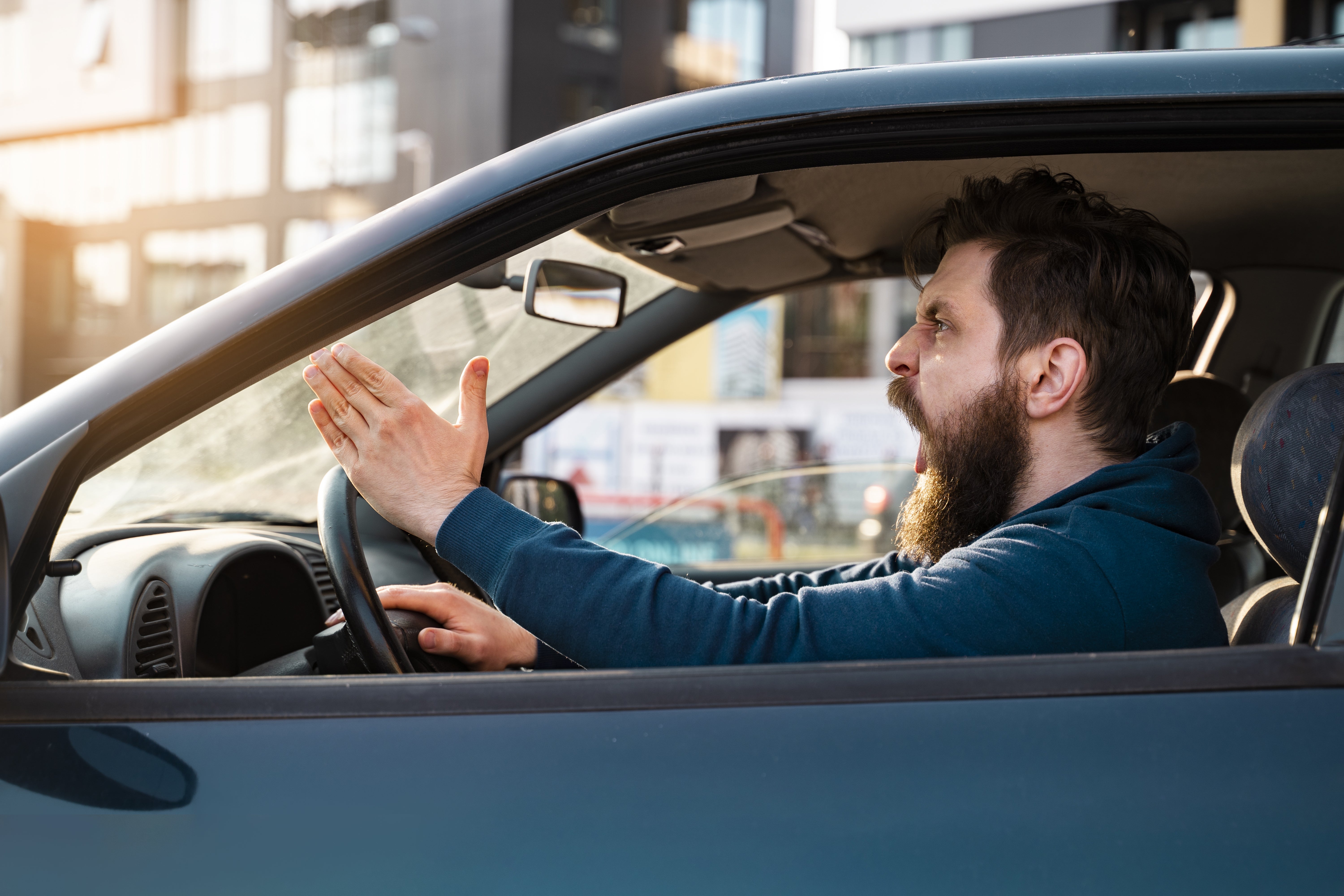 Man in a car expressing frustration while driving, gesturing with one hand, possibly indicating road rage or stress-related to traffic