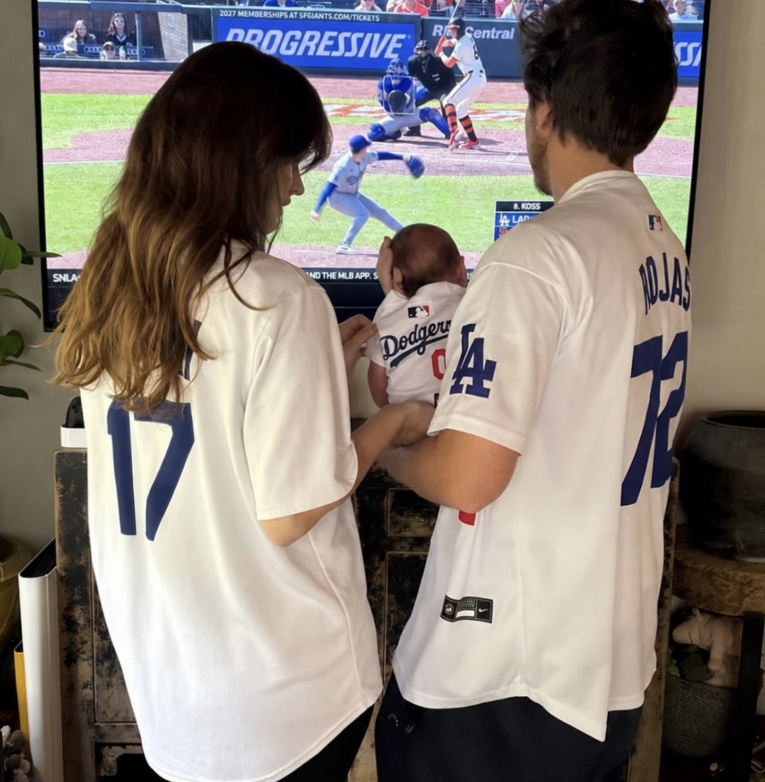 A family wearing Dodgers jerseys, including a baby, watches a baseball game on TV featuring the Dodgers and Mets