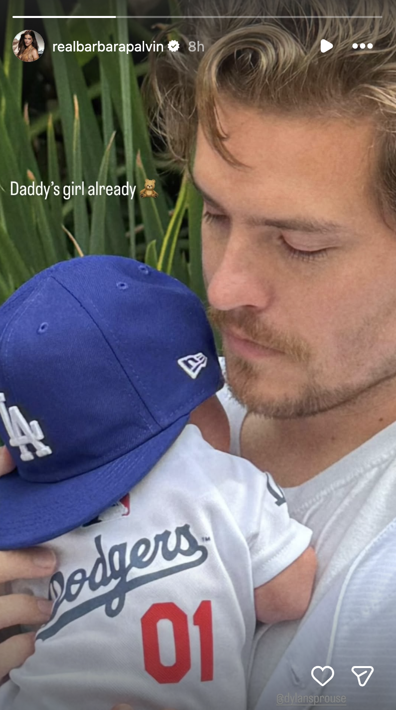 Person holds a baby wearing a baseball-themed outfit and cap, caption reads "Daddy's girl already."