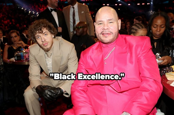 Two people sitting at an event; one in a beige suit, the other in a vibrant pink suit. Text reads "Black Excellence."