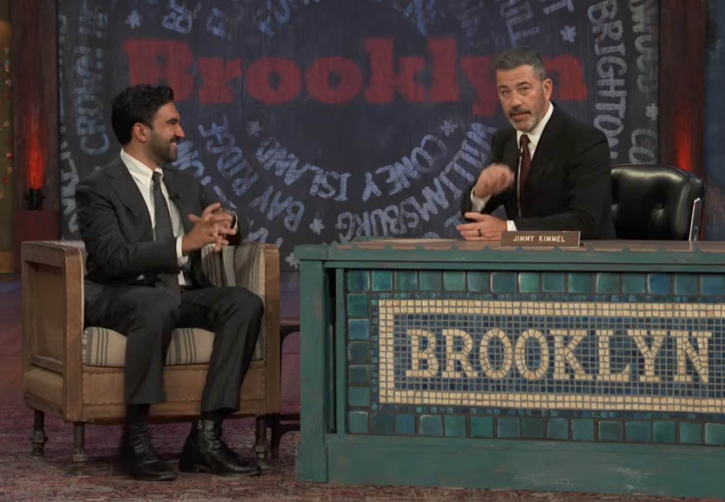 Talkshowvert intervjuer gjest. Vert i dress, gester mens du sitter bak et skrivebord merket 