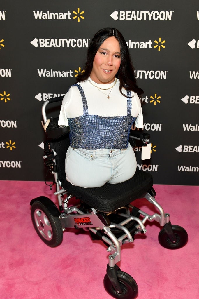 Person in a wheelchair on a pink carpet at an event, wearing a white top with a sequined overtop and light jeans. Sponsors' logos are in the background